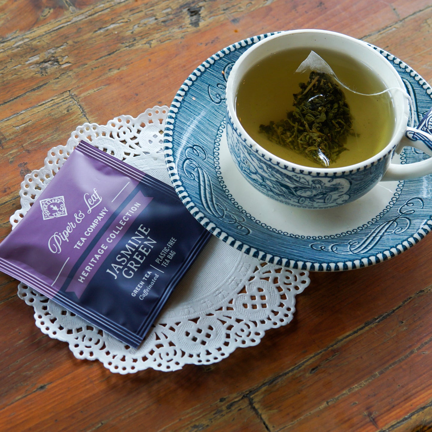 A blue and white teacup with a tea bag in hot water rests on a saucer; nearby on the wooden table are a paper doily and a Jasmine Green 50ct Individually Wrapped Tea Bag packet from Piper and Leaf Tea Co.’s Heritage Collection. The plastic-free tea bags from Piper and Leaf Tea Co. add an eco-friendly touch to your soothing tea experience.