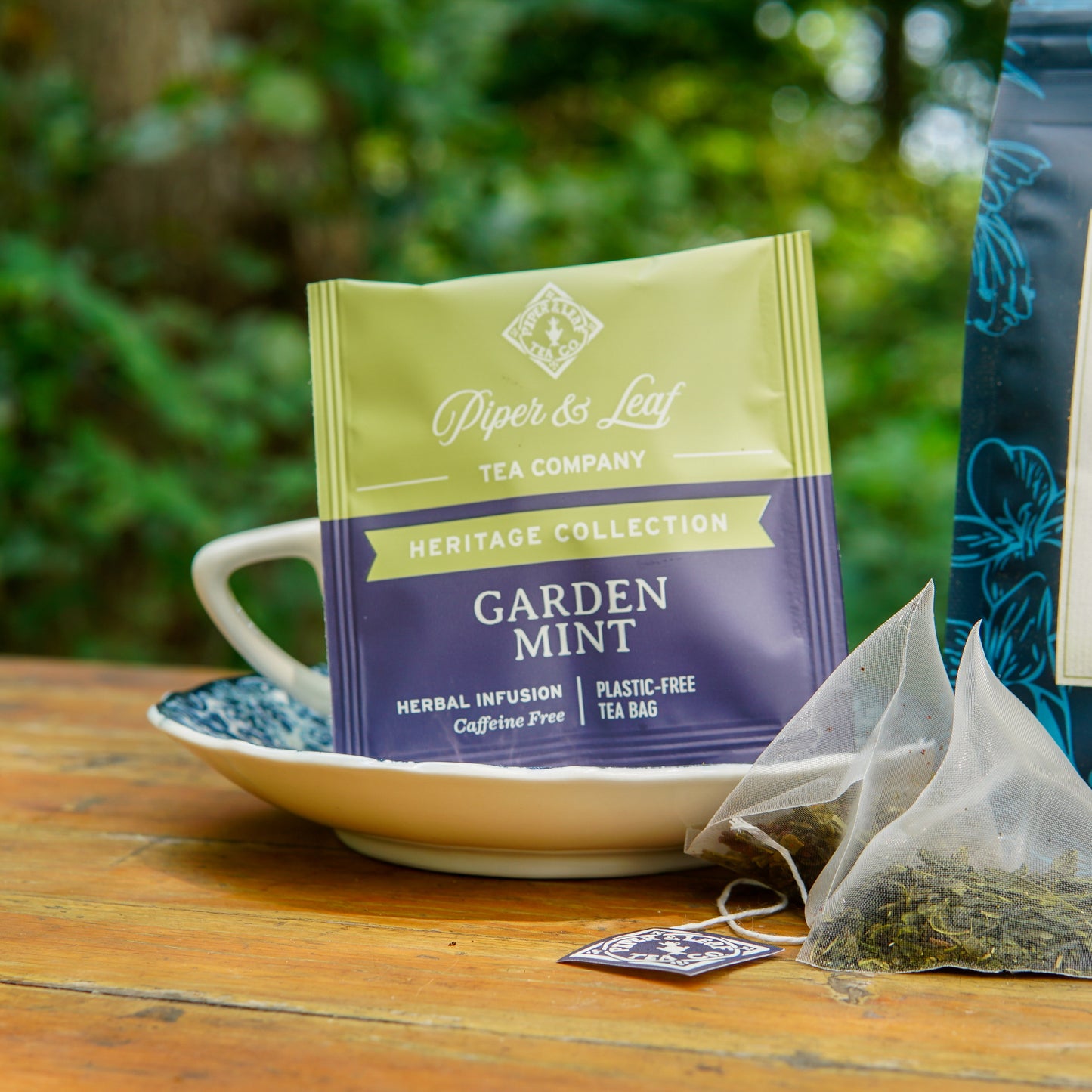 A Piper and Leaf Tea Co. Garden Mint 50ct Individually Wrapped Tea Bag rests on a teacup and saucer, with loose herbs scattered nearby atop a wooden outdoor table. This refreshing herbal blend is entirely caffeine freeāideal for peaceful garden relaxation, much like a classic peppermint chamomile tea.