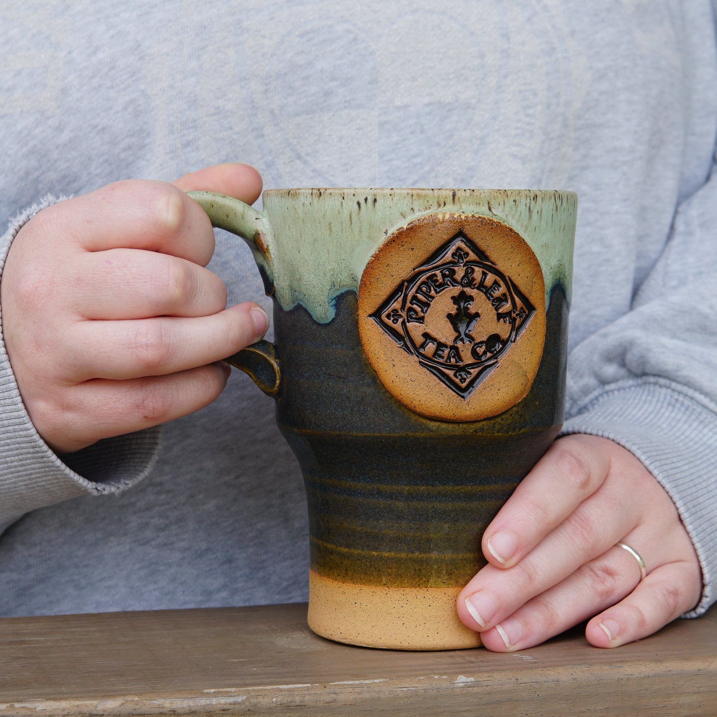 A person with light skin holds a large Pottery Travel Mug from Piper and Leaf Tea Co. while resting their hands on a wooden surface.