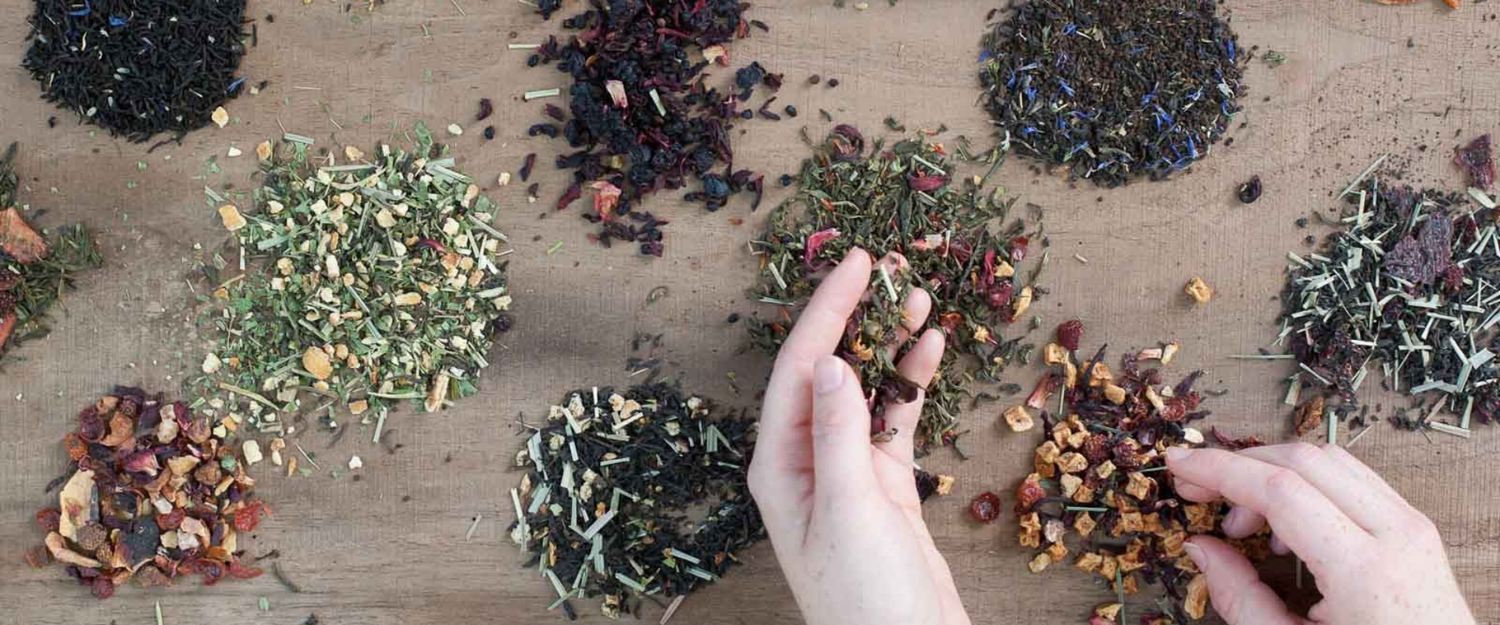 two hands placing different piper and leaf teas into little groupings on a wooden board 