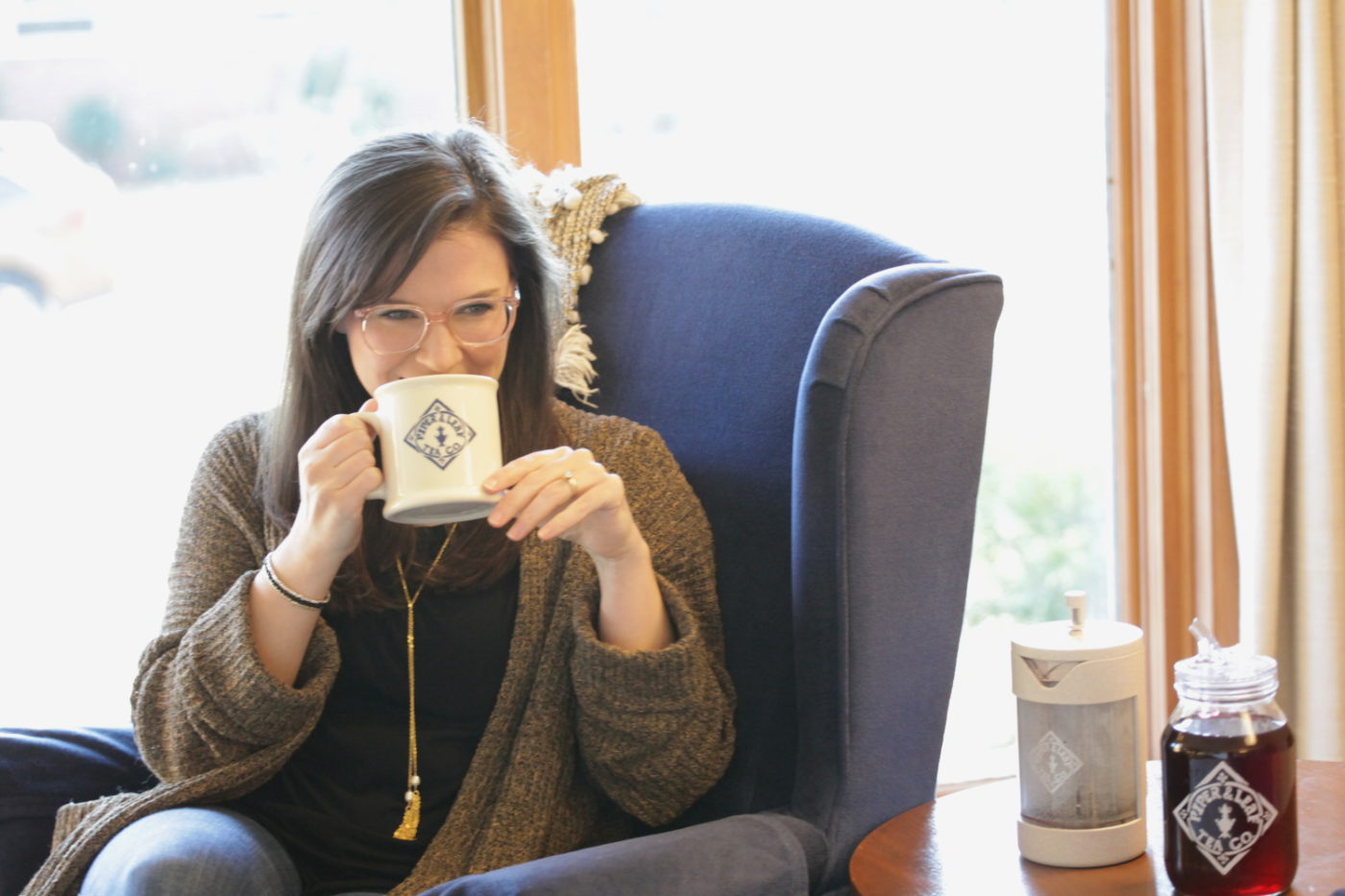 A woman in glasses sips hot tea from her Piper & Leaf mug beside her freshly brewed concentrated tea and her Piper Press