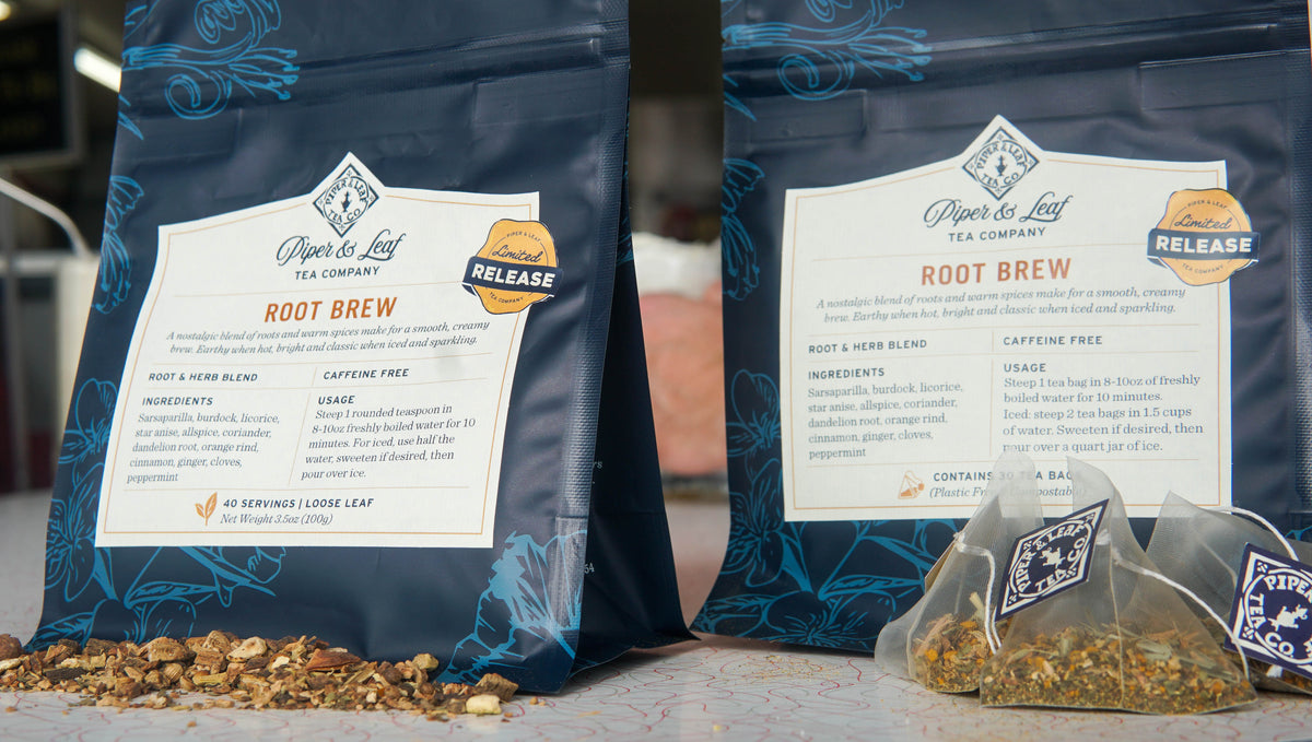 Two blue bags of Piper & Leaf Root Brew tea sit on a counter with loose tea ingredients scattered in front and three filled tea bags, showcasing product labels and ingredients.