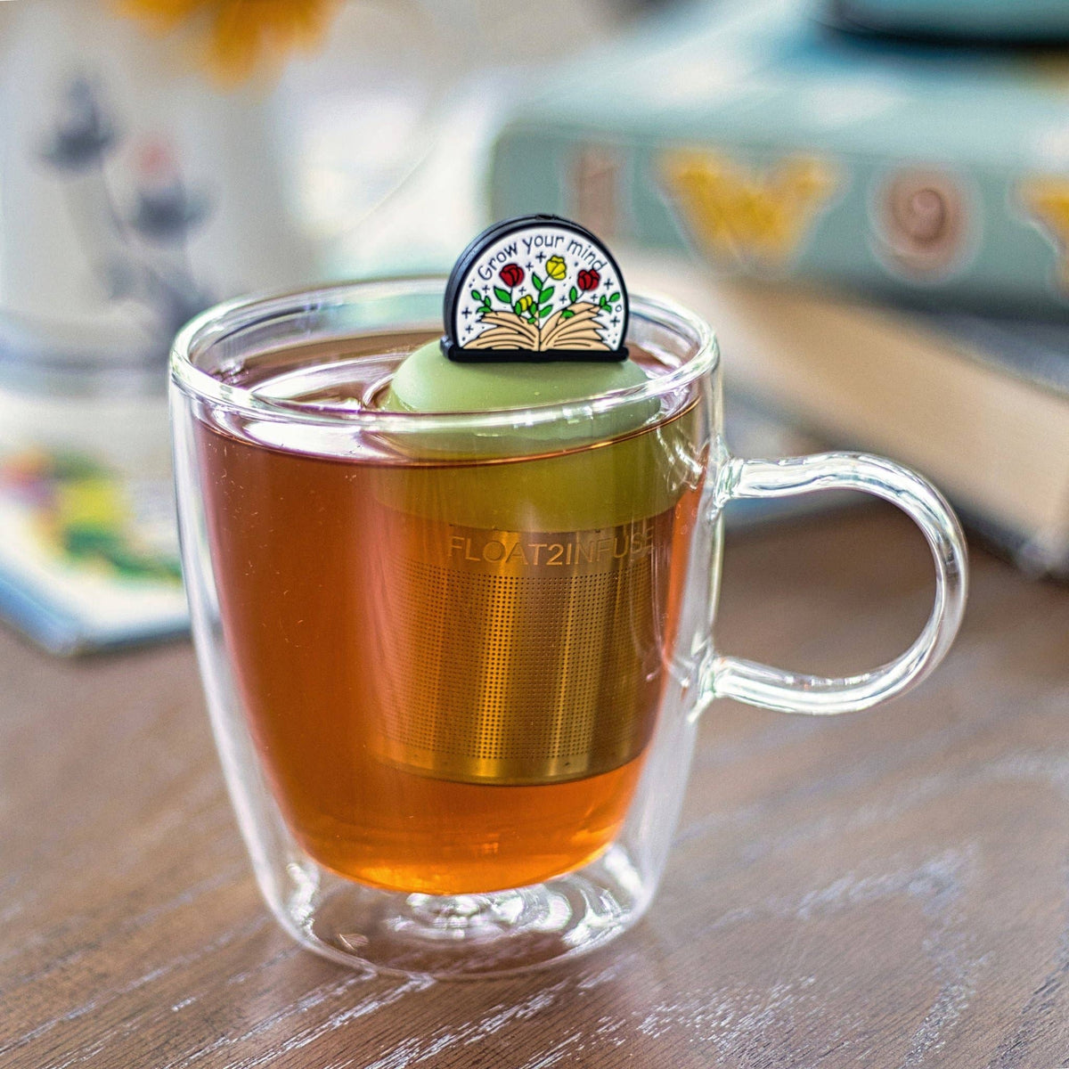 A clear glass mug of tea rests on a table alongside the Float2infuse Tea Infuser’s “Open Your Mind - Floating Tea Infuser” and a “Grow Your Mind” pin set on top. Blurred books and a floral mug in the background complete this cozy scene—perfect for any BookTok enthusiast.