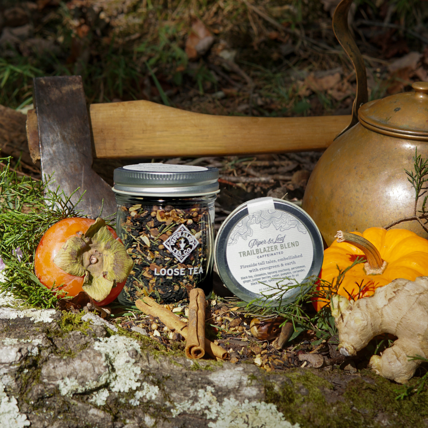 A Piper & Leaf Tea Co. Trailblazer Blend Glass Jar of Loose Leaf Tea (30 servings), labeled tea tin, persimmon, pumpkin, ginger, cinnamon sticks, axe, and brass kettle arranged outdoors on mossy ground.
