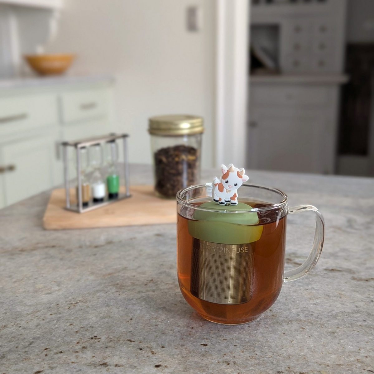 A glass mug of tea with the Float2infuse Tea Infuser’s Baby Goat - Floating Tea Infuser bobbing inside, its small goat figurine perched on top, sits on a kitchen counter surrounded by jars and spices.