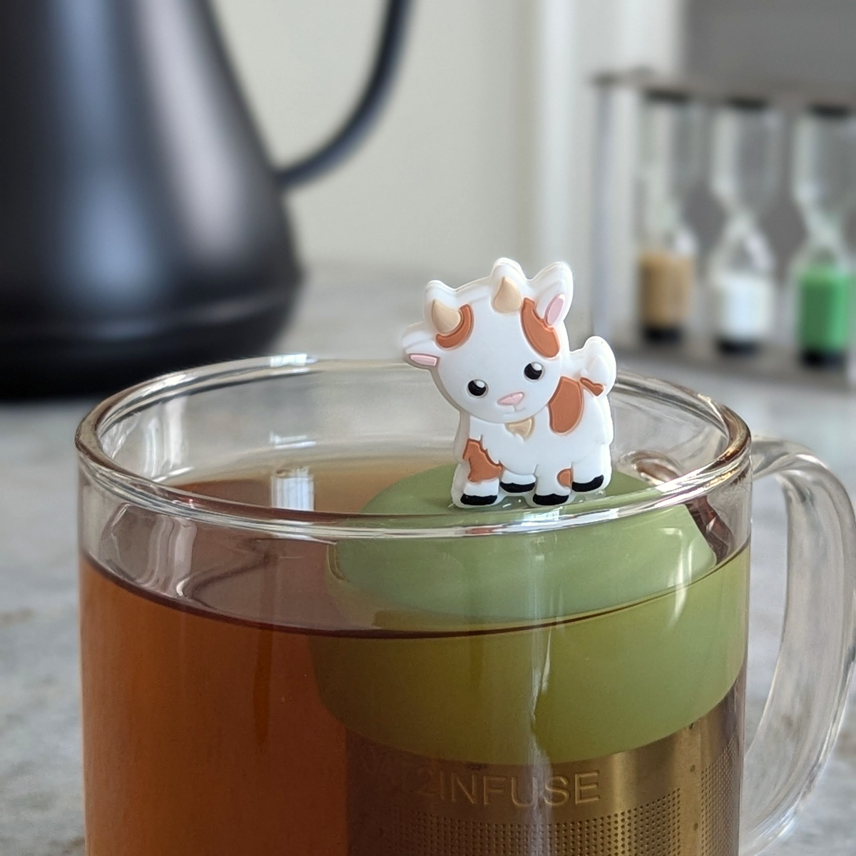 A glass mug of tea with the Float2infuse Tea Infuser’s “Baby Goat” floating loose-leaf steeper and a delightful baby goat figurine perched on top. A kettle and hourglass timers are softly blurred in the background, bringing focus to this charming tea accessory.