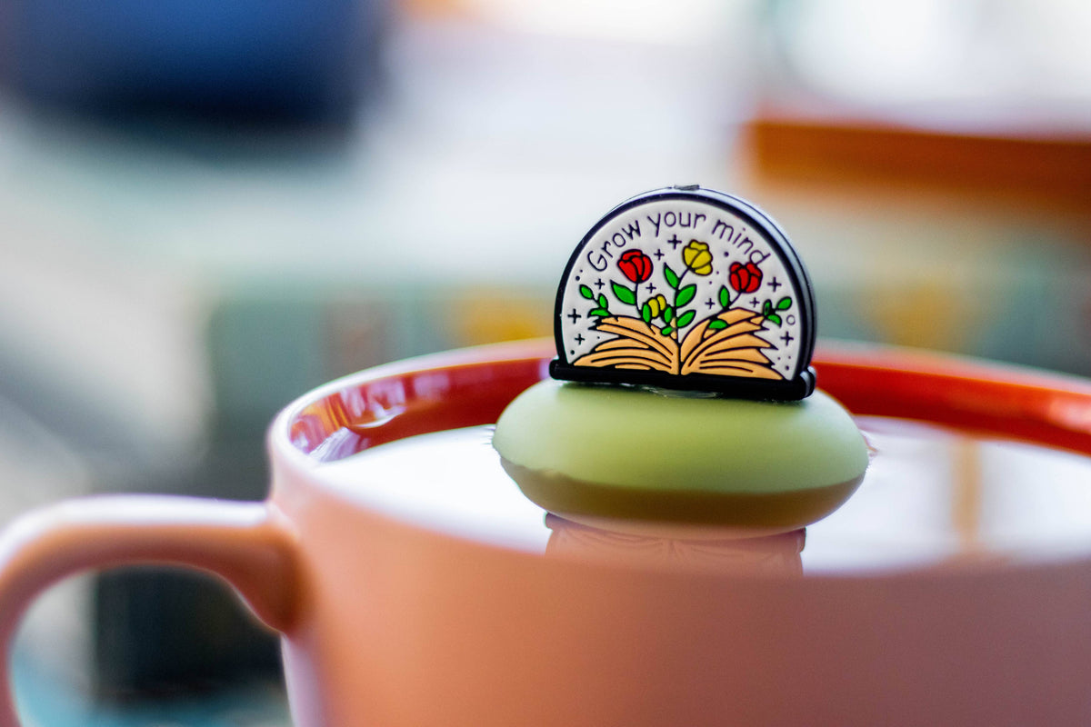 The Float2infuse Tea Infuser’s “Open Your Mind - Floating Tea Infuser,” featuring charming plant and book artwork and the phrase “Grow your mind,” rests atop a green, floating tea infuser in a pink mug—an ideal loose-leaf tea accessory for BookTok fans.