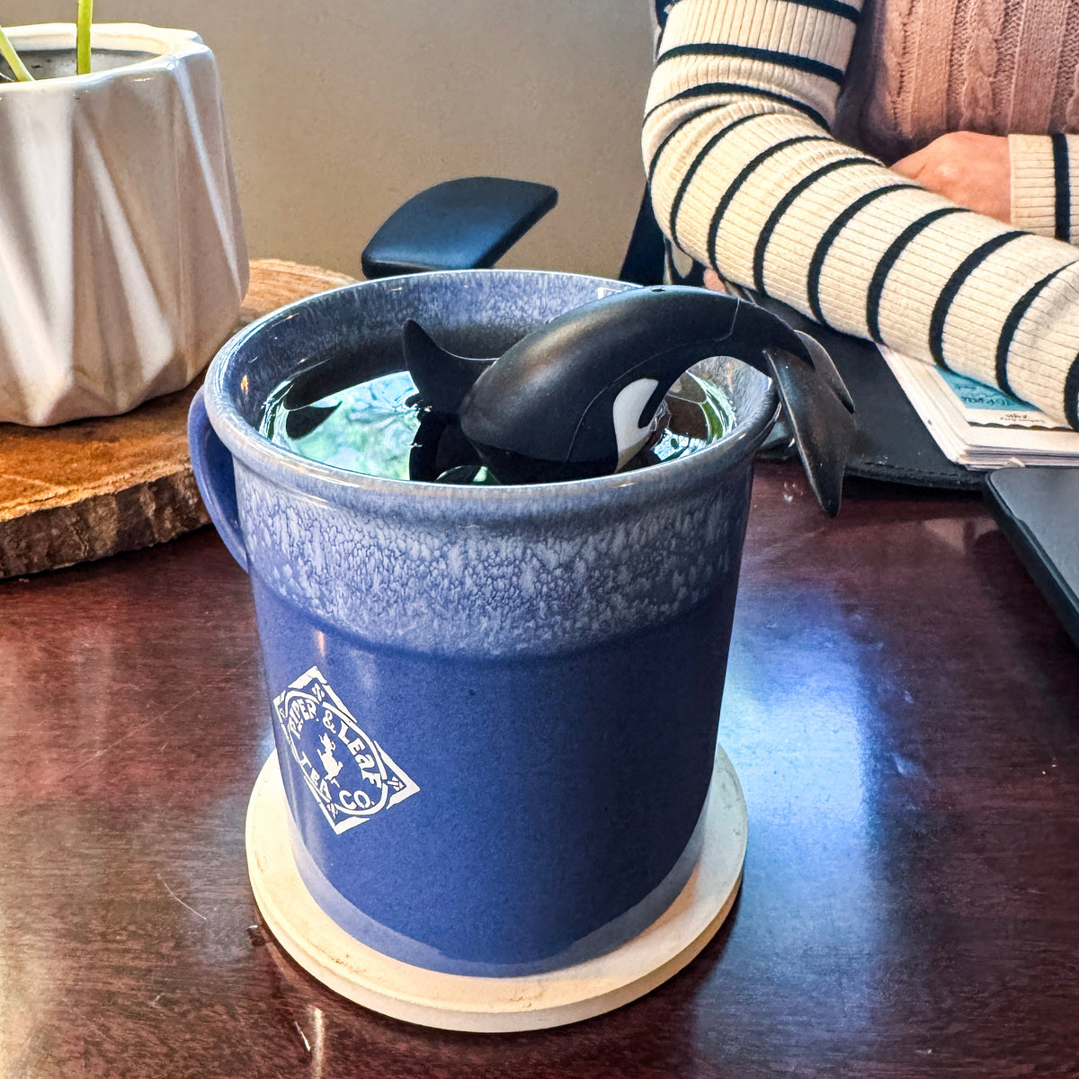 A Piper & Leaf Tea Co. Steep Dive Infuser, shaped like a black and white orca and safe for dishwashers, rests in a blue ceramic mug filled with tea on a wooden desk beside a notepad and someone in a striped sweater.