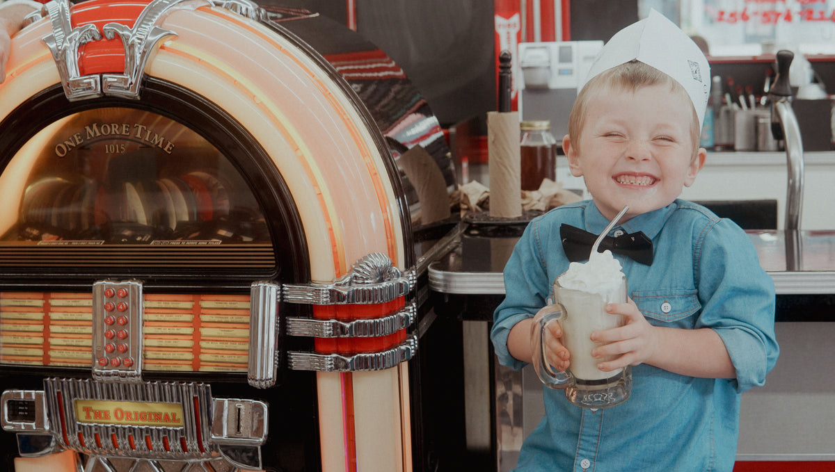 A young boy in a paper hat and bow tie smiles while holding a Piper and Leaf Tea Co. Root Brew Kit next to a vintage jukebox in a retro diner.