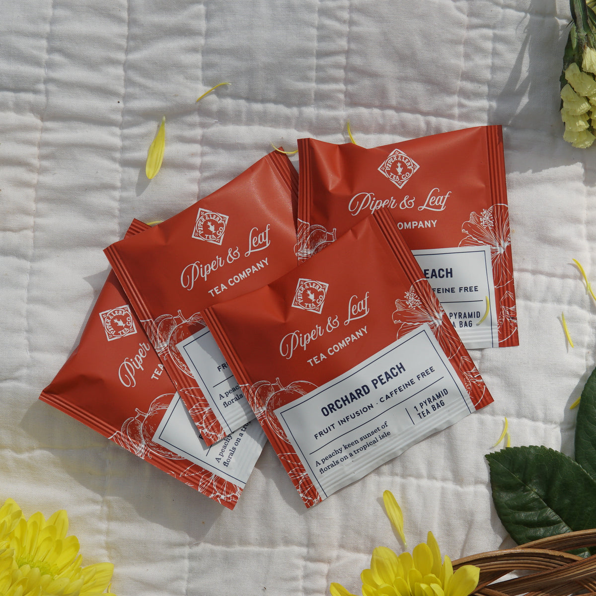 Sixteen individually wrapped Piper & Leaf Tea Co. Mother's Day Special Tea Sampler bags are displayed on a white quilt with yellow petals and green leaves—an ideal herbal tea gift or relaxing Mother's Day treat.
