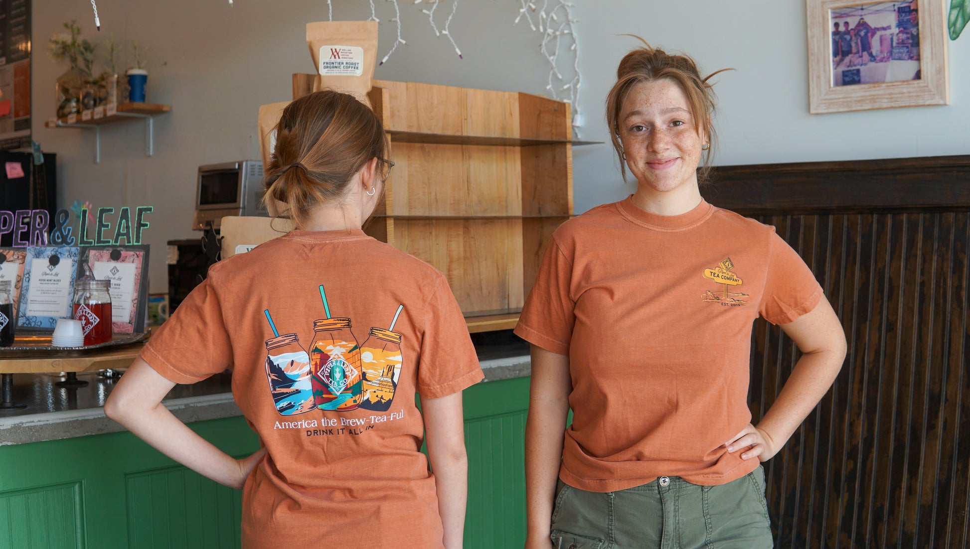 Two young women in orange Westward Adventure Shirts by Piper & Leaf Tea Co. stand in a cafe; one smiles at the camera, while the other displays the back featuring colorful drink illustrations and fun text.