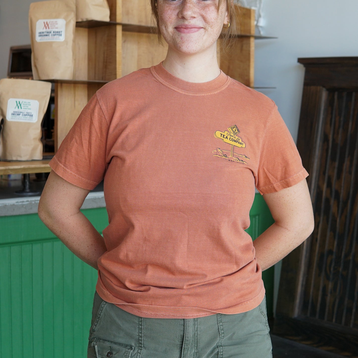A person wearing the limited edition Westward Adventure Shirt by Piper & Leaf Tea Co. in orange and green pants smiles indoors with hands behind their back. Shelves filled with coffee bags appear in the background.