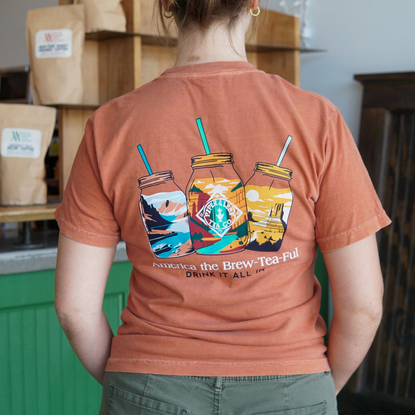 A person wearing the Westward Adventure Shirt by Piper & Leaf Tea Co., a limited edition orange tee with three mason jars and "America the Brew-Tea-Ful, Drink It All In" illustrated on the back.