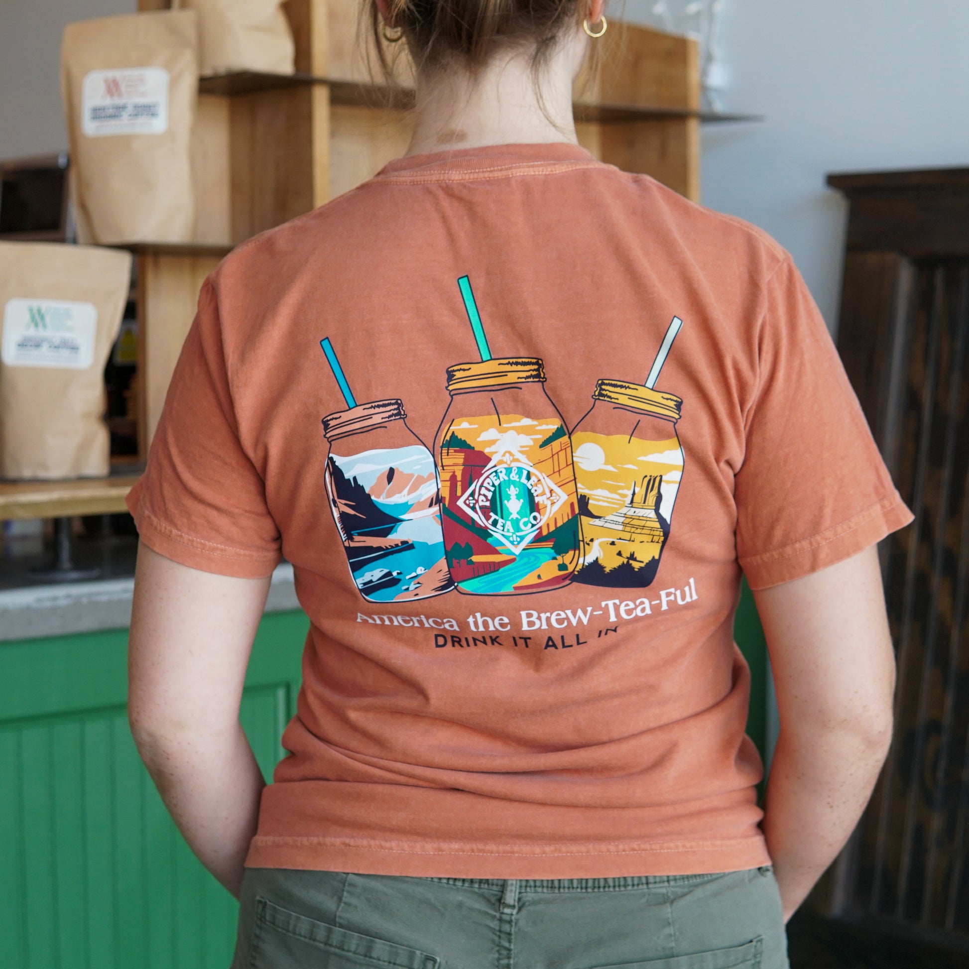 A person wearing the Westward Adventure Shirt by Piper & Leaf Tea Co., a limited edition orange tee with three mason jars and "America the Brew-Tea-Ful, Drink It All In" illustrated on the back.
