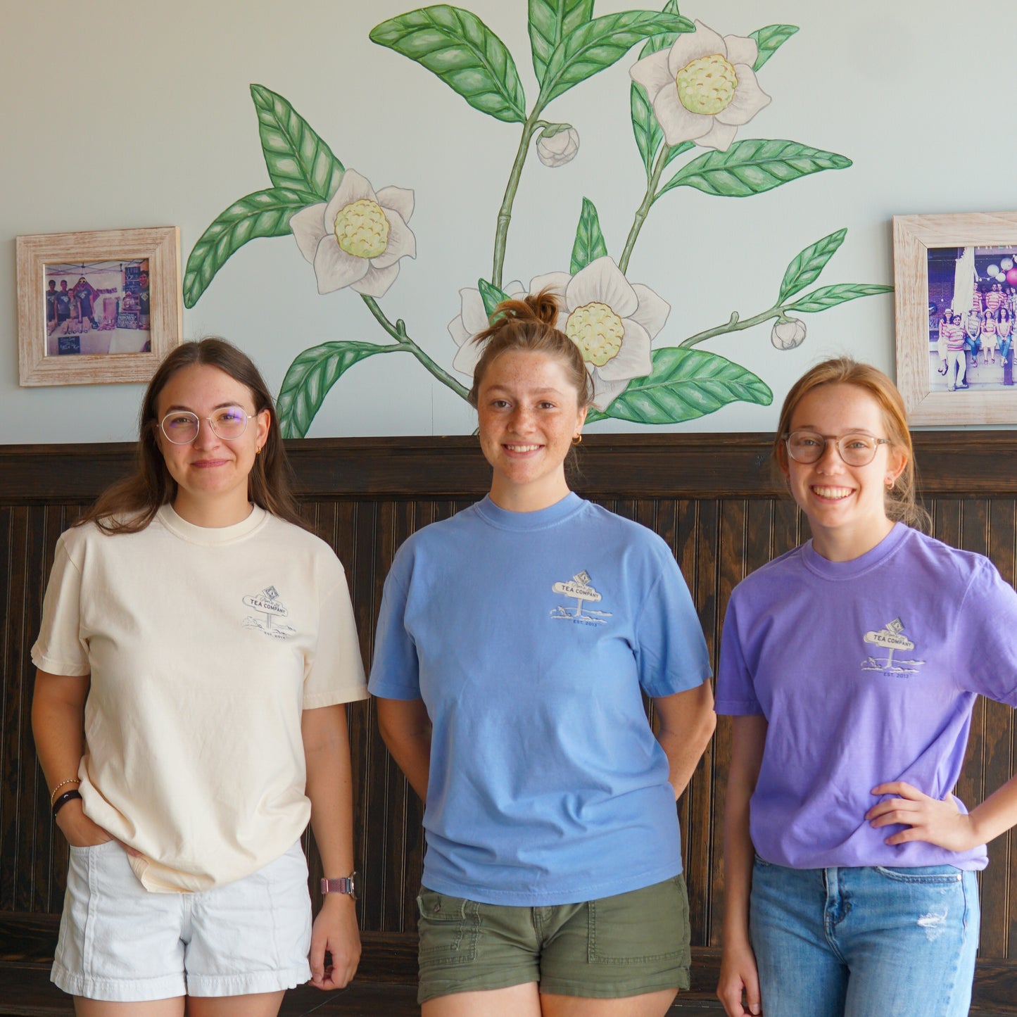Three young women stand indoors, smiling in front of a leafy wall mural while wearing Nor'Easter Adventure T-shirts by Piper and Leaf Tea Co., paired with casual shorts.