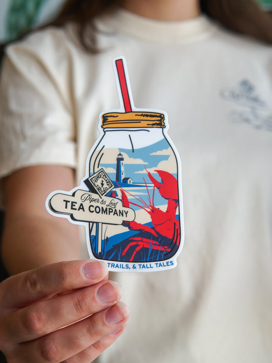 A person holds the Nor'Easter Adventure Sticker by Piper and Leaf Tea Co., featuring a mason jar of tea with a red straw, a lobster, a "Tea Company" tag, and a lighthouse in the background.