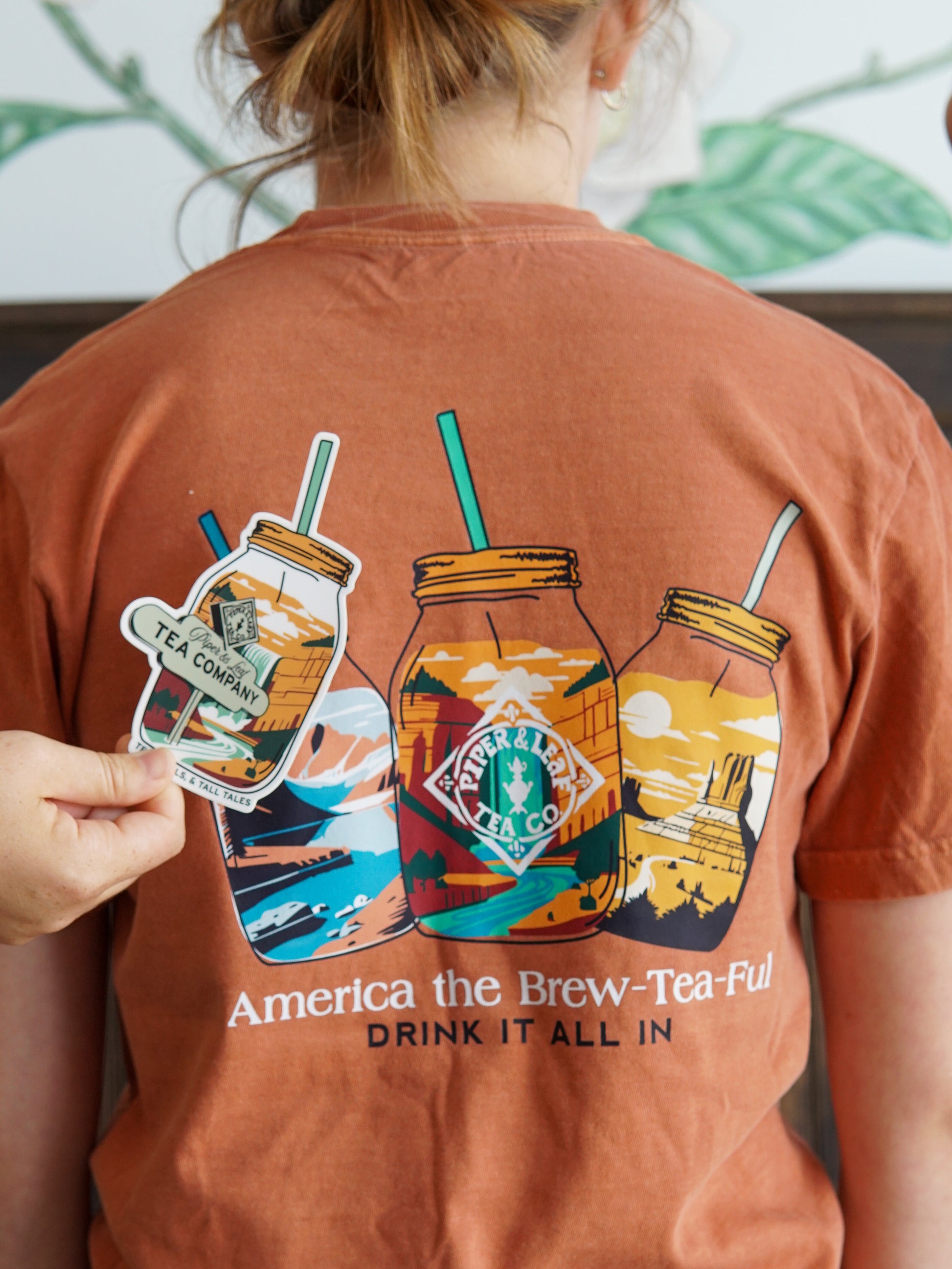 A person wears the limited edition Piper & Leaf Tea Co. “Westward Adventure Shirt” featuring illustrated mason jars and outdoor scenes, holding a matching sticker. Text: "America the Brew-Tea-Ful. Drink it all in." For adventure tee fans.