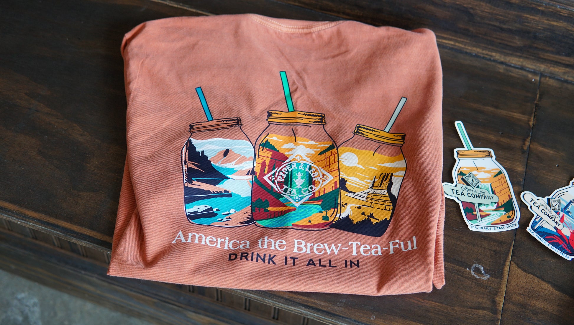 The Westward Adventure Shirt by Piper & Leaf Tea Co. is an orange tee with scenic mason jar designs, “Franklin Tea Co.,” and “America the Brew-Tea-Ful DRINK IT ALL IN.” This limited edition comes with fun stickers beside the shirt.