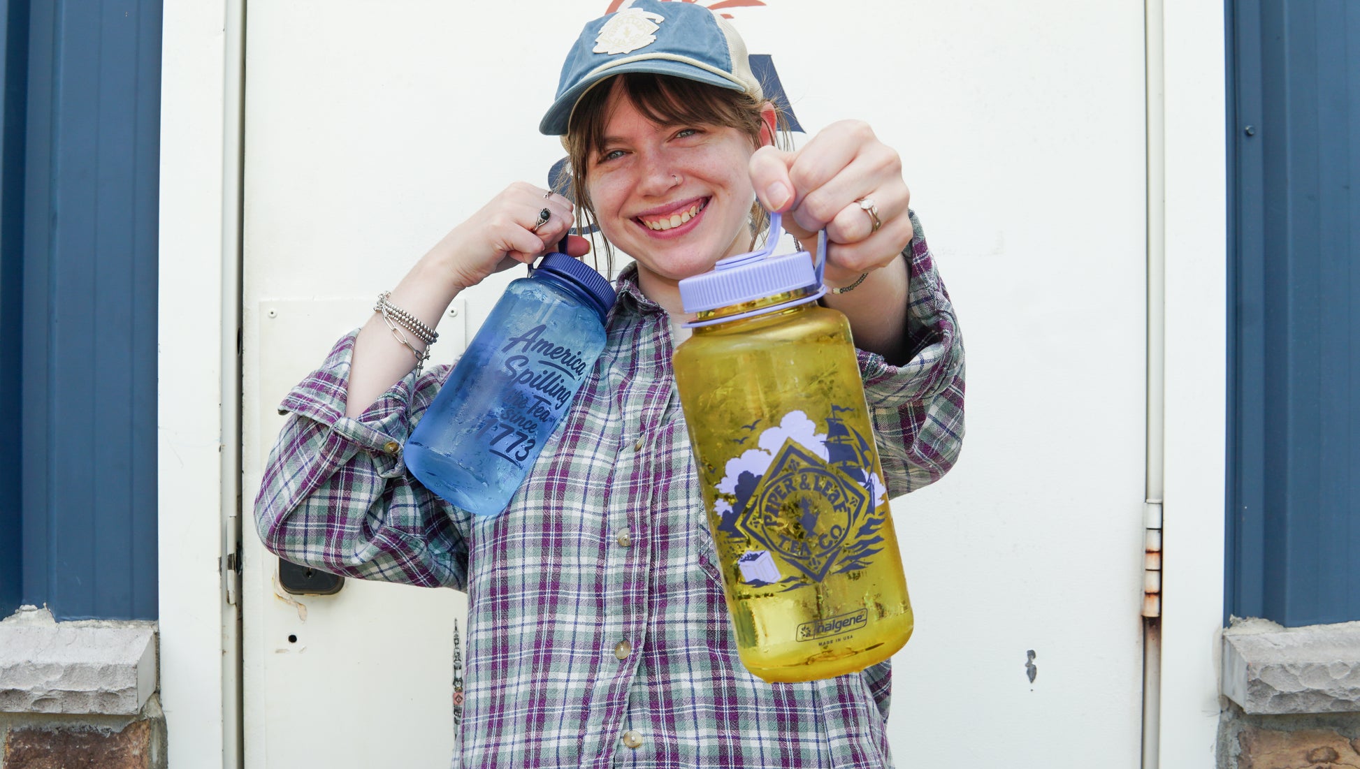 A person in a plaid shirt and cap smiles while holding up a blue Spilling the Tea Nalgene and a yellow Spilling the Tea Nalgene by Piper and Leaf Tea Co.