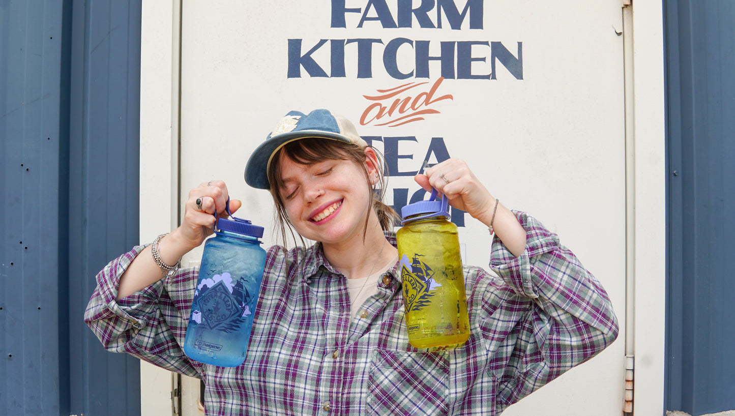 A person in a plaid shirt and cap smiles, holding two large Spilling the Tea Nalgene bottles by Piper and Leaf Tea Co.—one blue, one yellow—in front of a sign that says "FARM KITCHEN and SEA.