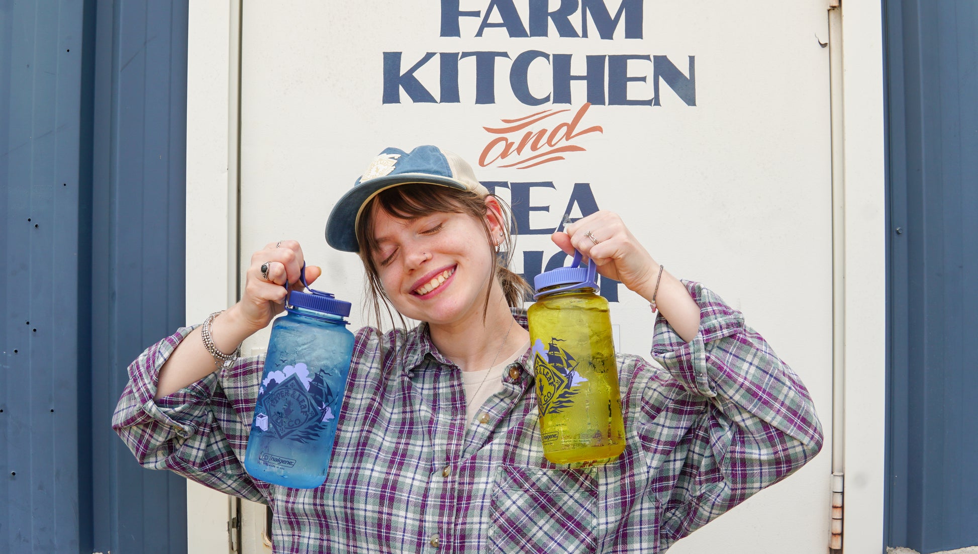 A person in a plaid shirt and cap smiles, holding two large Spilling the Tea Nalgene bottles by Piper and Leaf Tea Co.—one blue, one yellow—in front of a sign that says "FARM KITCHEN and SEA.