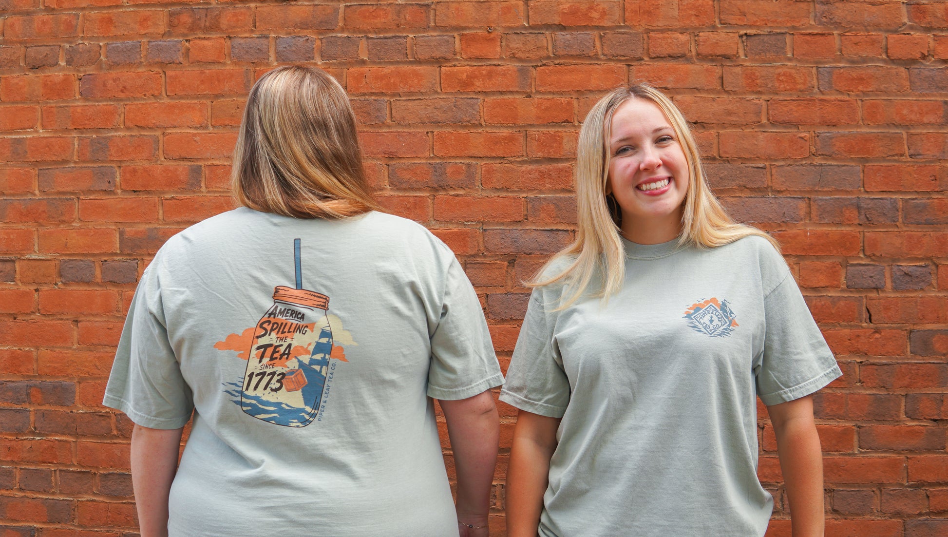 Two women pose against a brick wall. One smiles at the camera while the other shows off the back of her Piper and Leaf Tea Co. Spilling the Tea Shirt, which features a mason jar and the text "Spilling Tea 1773.