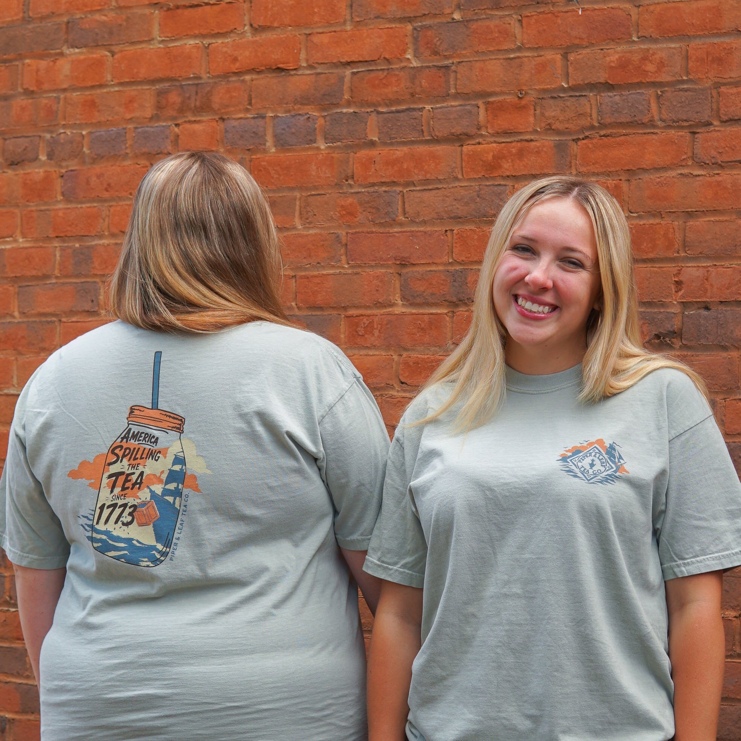 Two women stand by a brick wall; one smiles at the camera while the other displays the back of her Piper and Leaf Tea Co. "Spilling the Tea Shirt" featuring a “Spilling Tea 1773” graphic.