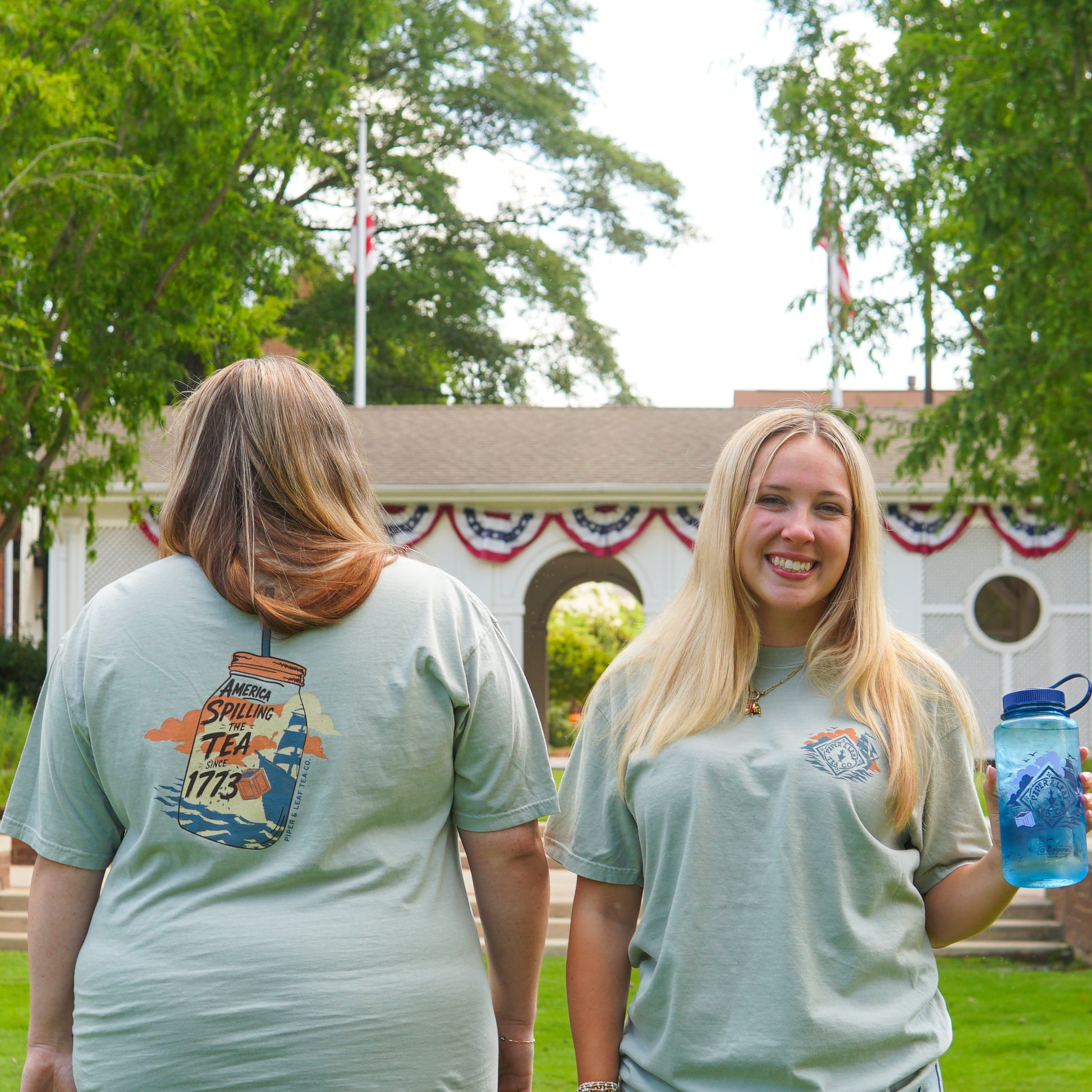 Two women are outdoors; one smiles at the camera holding a blue water bottle, while the other displays the back of her Piper and Leaf Tea Co. "Spilling the Tea Shirt" featuring a colorful printed design.