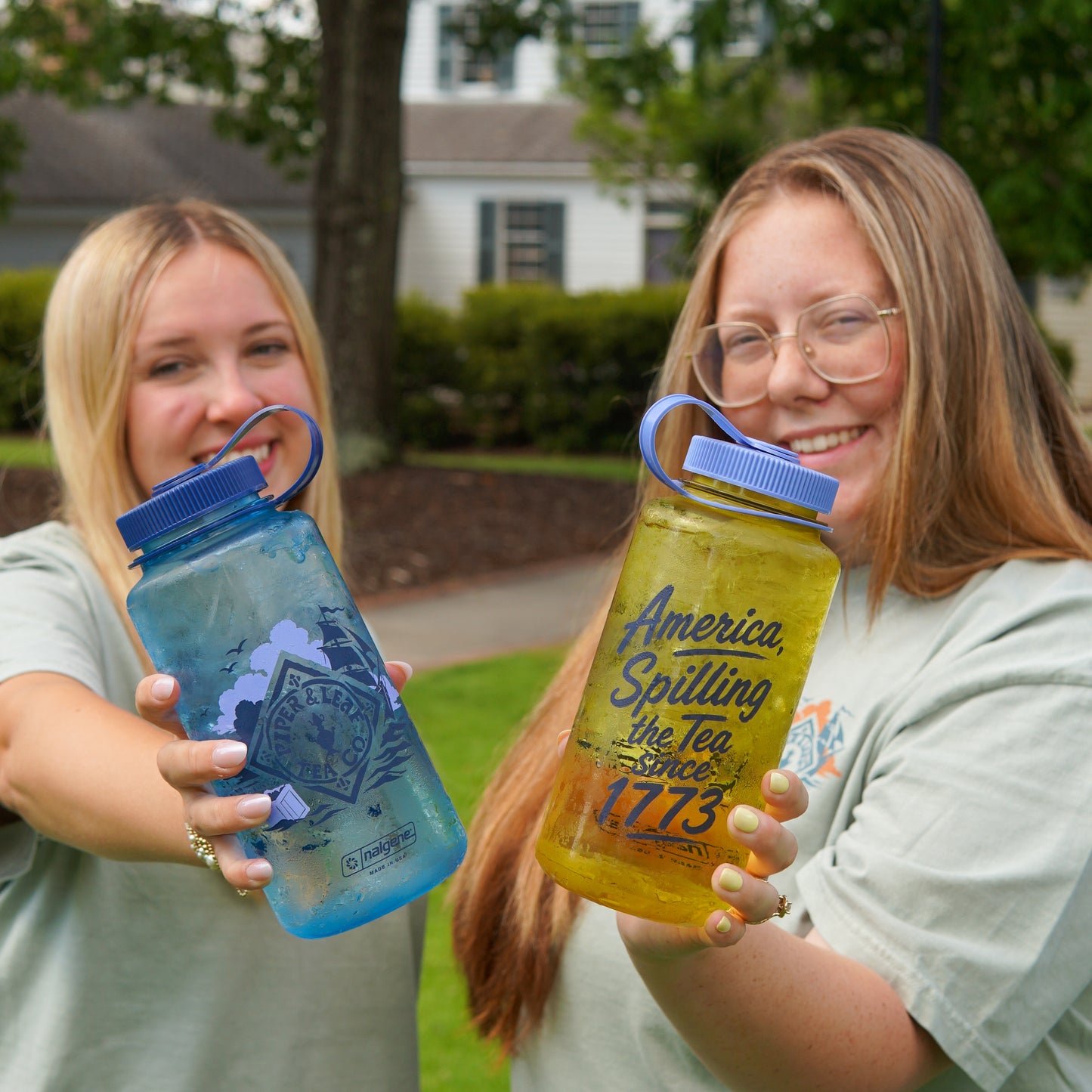 Two young women outdoors smile at the camera, each holding a large water bottle featuring printed tea-spilling designs and "1773," promoting the Spilling the Tea Shirt by Piper and Leaf Tea Co.
