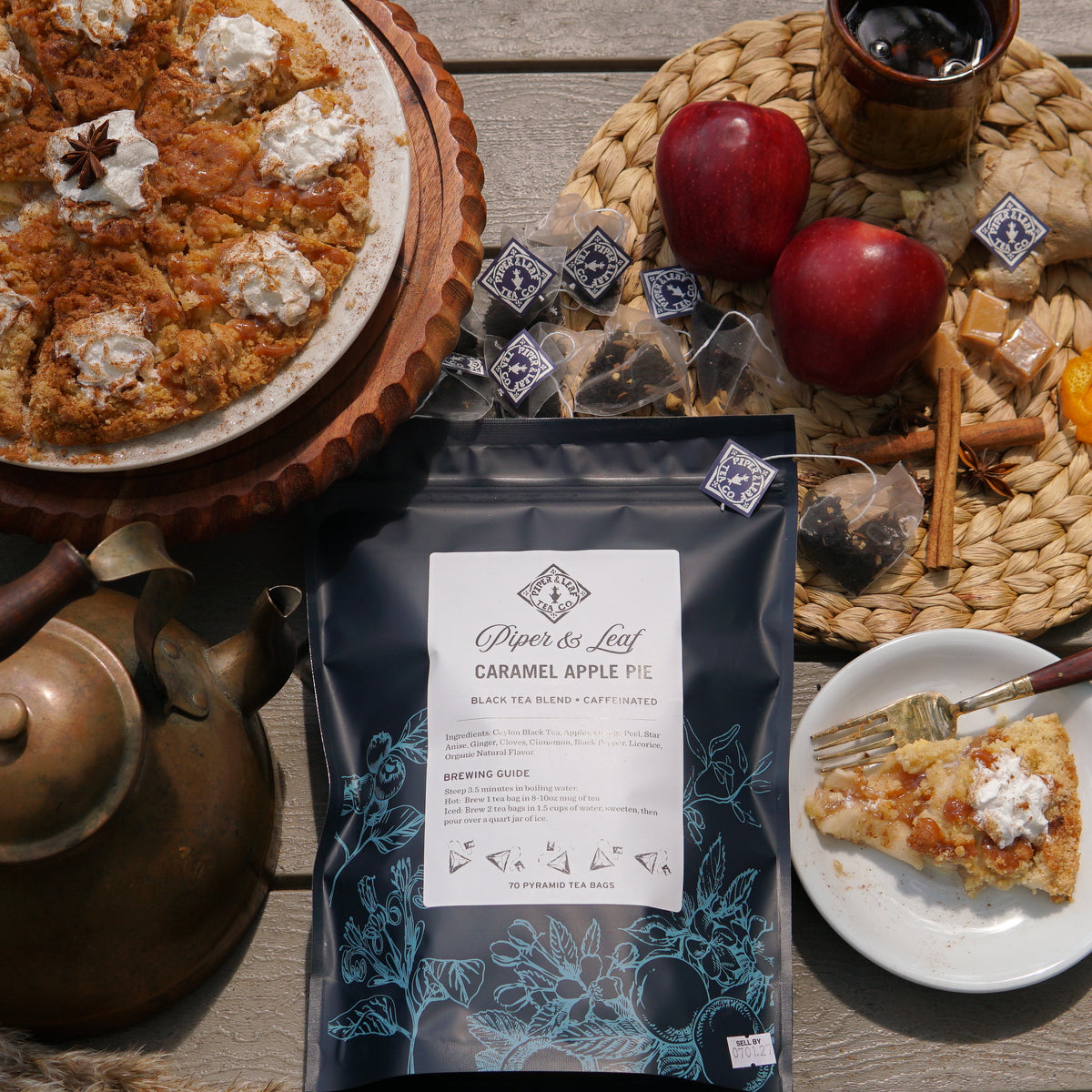 A caramel apple pie with whipped cream, a box of Piper & Leaf Tea Co. Caramel Apple Pie - 70ct Tea Bags, glass of tea, teapot, and wrapped tea bags are displayed on a wooden table.