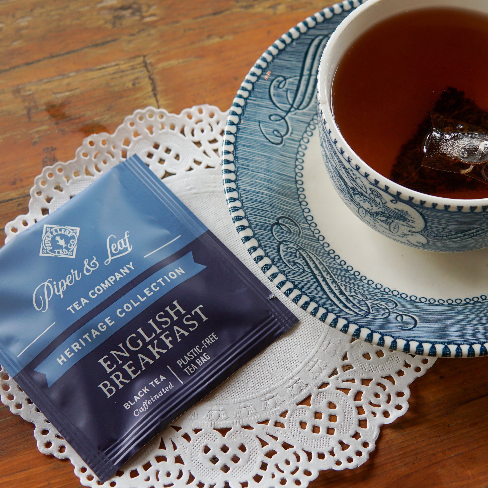 A cup of tea rests on a blue-patterned saucer beside a packet of Piper and Leaf Tea Co. English Breakfast 50ct Individually Wrapped Tea Bags, all arranged on a lace doily atop a wooden table.