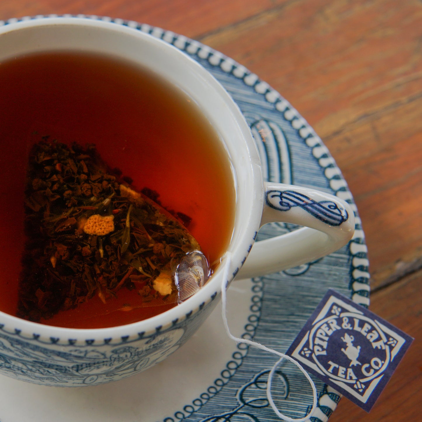 A cup of Earl Grey tea, brewed with a triangular bag from the Earl Grey 30ct Tea Bags by Piper and Leaf Tea Co., sits on a blue and white patterned saucer. The bag’s label, bearing the Piper and Leaf Tea Co. name, hangs over the edge of the cup.