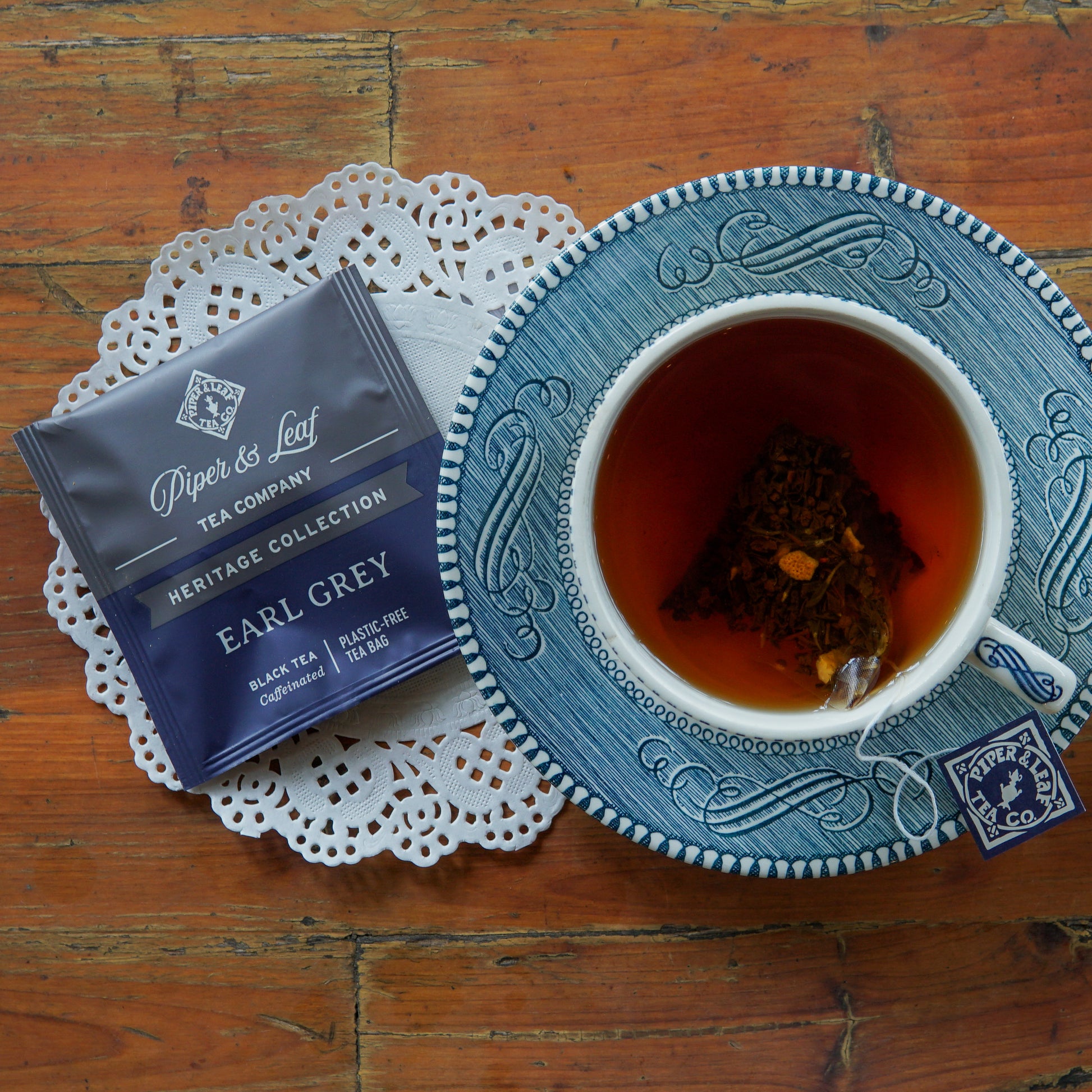 A cup of Earl Grey tea and a tea bag rest on a blue saucer, beside a Piper and Leaf Tea Co. Earl Grey 50ct Individually Wrapped Tea Bags packet from the Heritage Collection, displayed on a doily atop a wooden table.