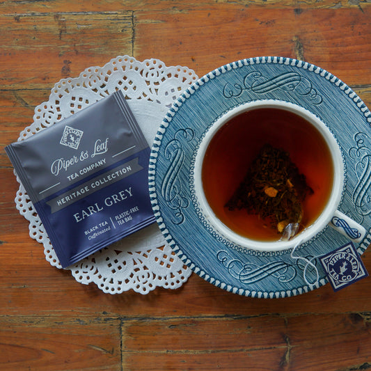 A cup of Earl Grey tea and a tea bag rest on a blue saucer, beside a Piper and Leaf Tea Co. Earl Grey 50ct Individually Wrapped Tea Bags packet from the Heritage Collection, displayed on a doily atop a wooden table.