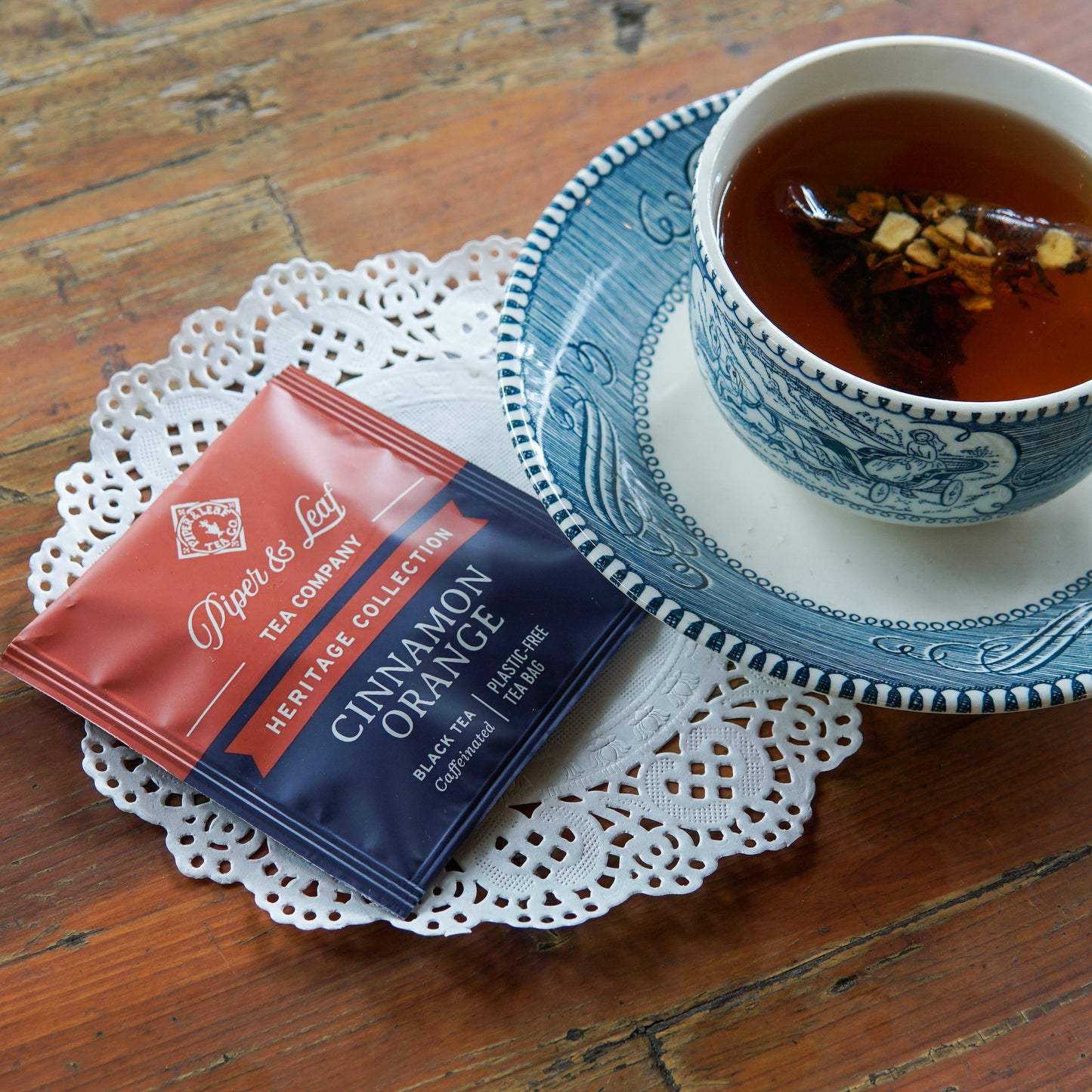 A blue and white teacup filled with tea sits on a saucer beside a doily and Piper and Leaf Tea Co. Cinnamon Orange 50ct Individually Wrapped Tea Bags on a wooden table.