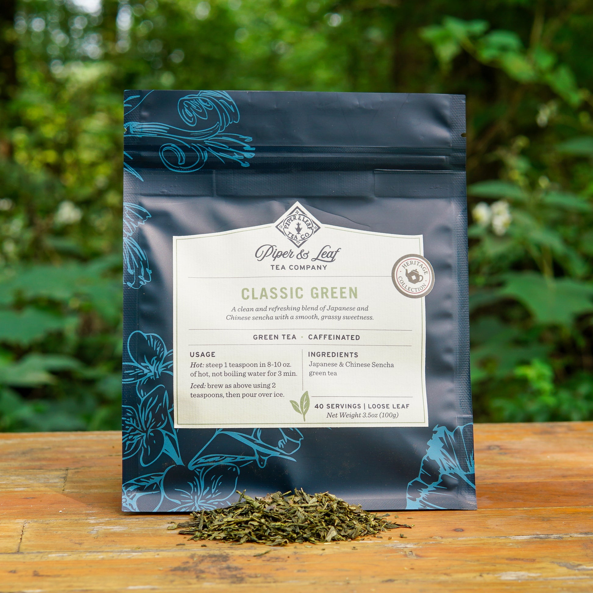 A package of Piper and Leaf Tea Co. Classic Green 40 Serving Loose Leaf sits on a wooden surface outdoors, with a small pile of sencha green tea leaves in front of it and greenery in the background.