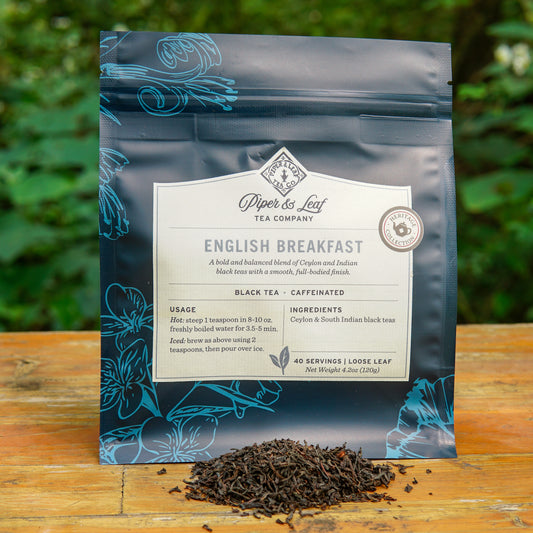 A blue bag of Piper and Leaf Tea Co. English Breakfast 40 Serving Loose Leaf is displayed outdoors, with a small pile of Ceylon black tea leaves arranged in front of the bag.