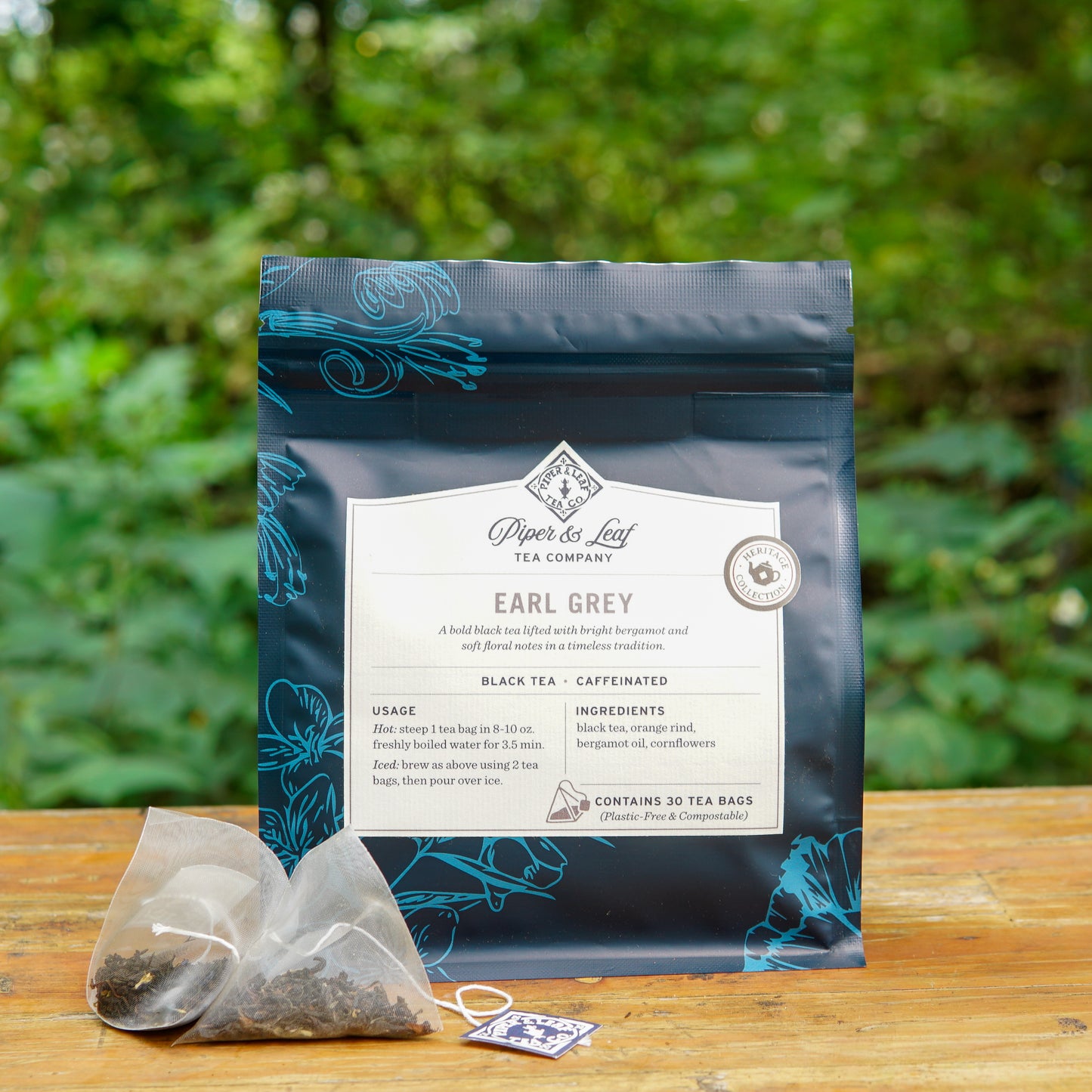 A bag of Piper and Leaf Tea Co. Earl Grey 30ct Tea Bags from the Heritage Collection rests on a wooden surface outdoors beside a pyramid tea bag, with lush greenery softly blurred in the background.