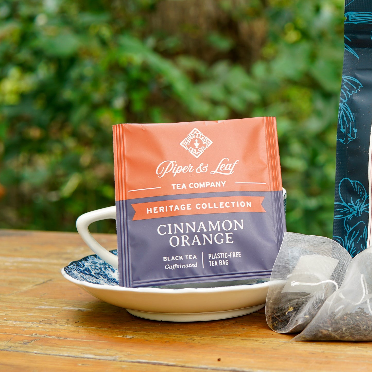 A Piper and Leaf Tea Co. Cinnamon Orange 50ct Individually Wrapped Tea Bag from the Heritage Collection rests on a white and blue saucer, accompanied by a tea cup and another black tea bag, all arranged on a wooden outdoor table.