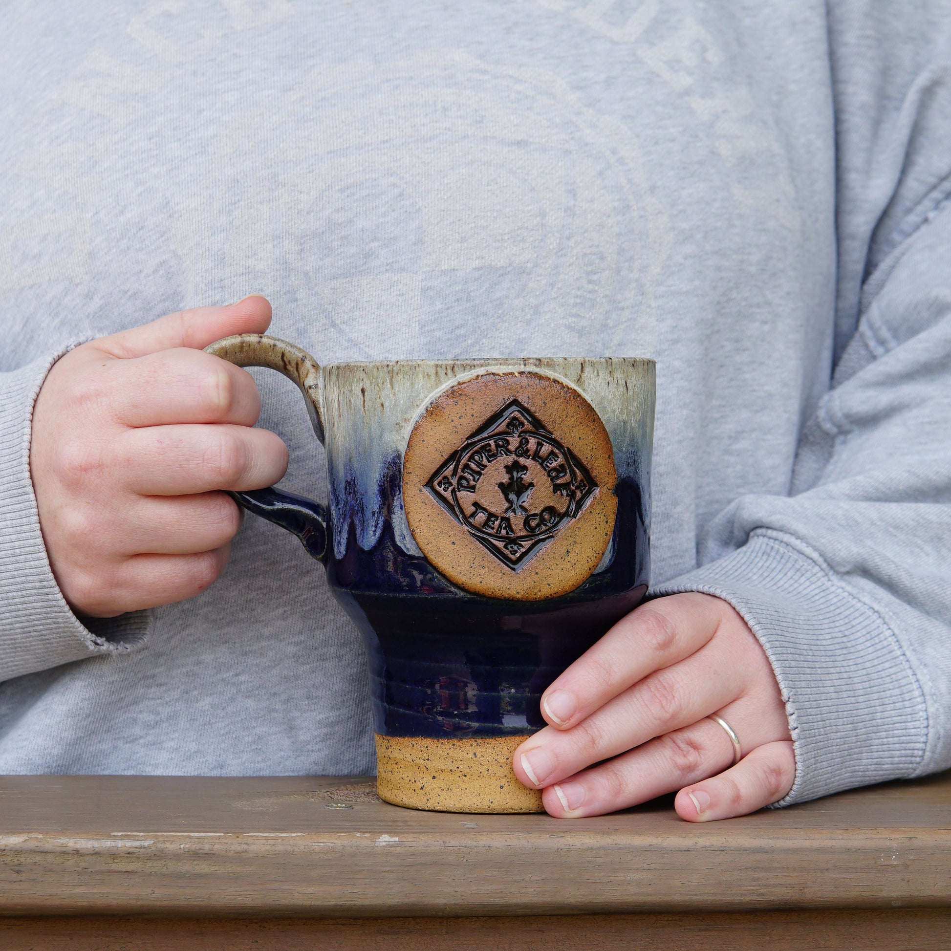 Person in a light gray sweatshirt holds a large Pottery Travel Mug featuring the Piper and Leaf Tea Co. logo, resting on a wooden surface.
