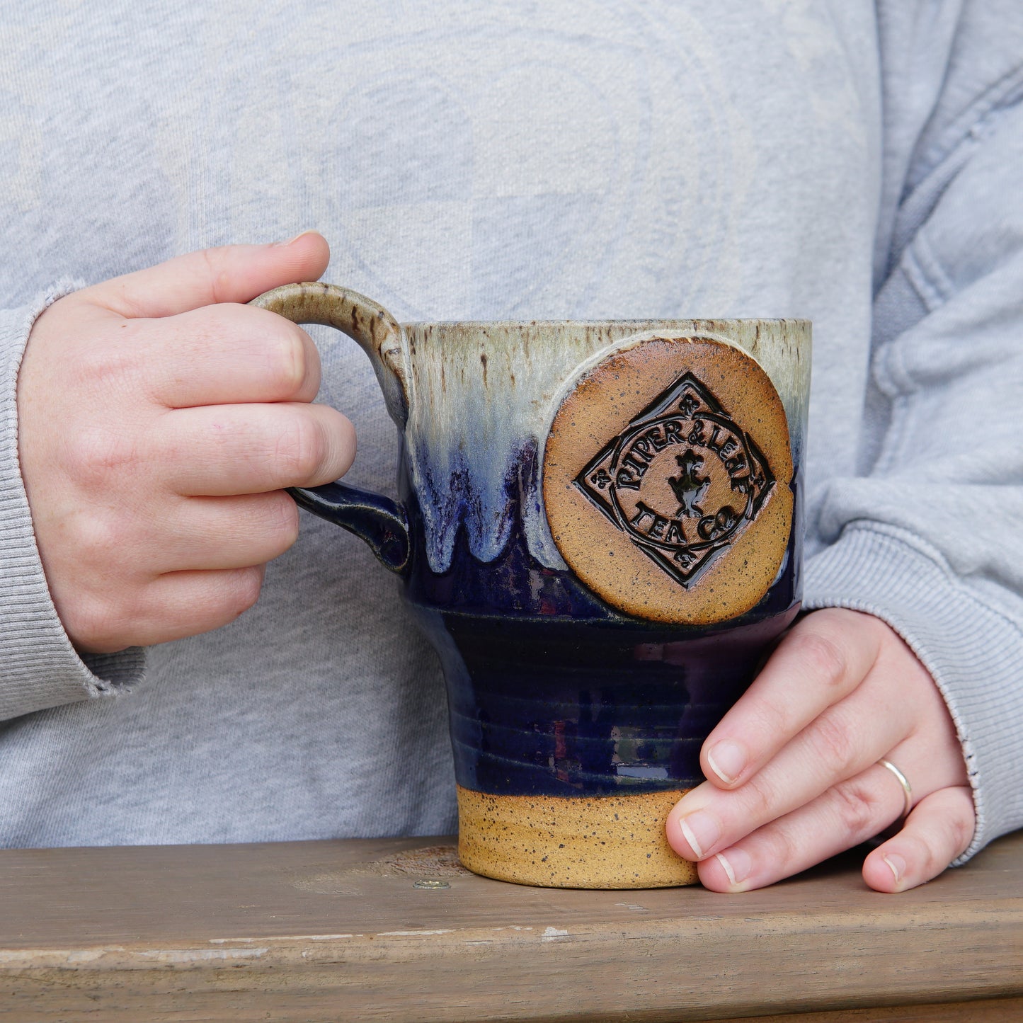 A person holding a Pottery Travel Mug with a “Piper and Leaf Tea Co.” logo, standing at a wooden surface, wearing a light gray sweatshirt.