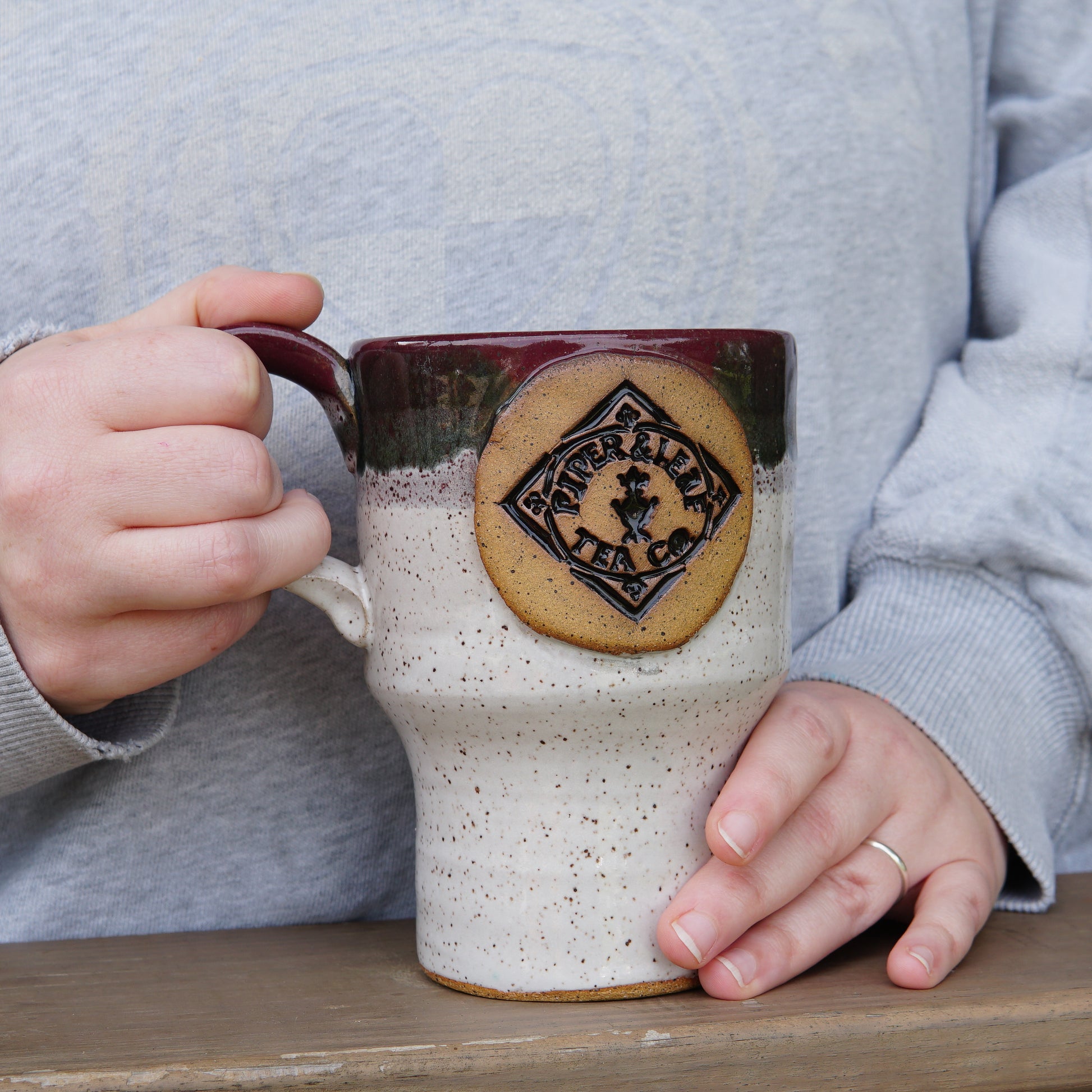 A person holding a Pottery Travel Mug with a “Piper and Leaf Tea Co.” logo, wearing a light gray sweatshirt and a silver ring, standing by a wooden surface.