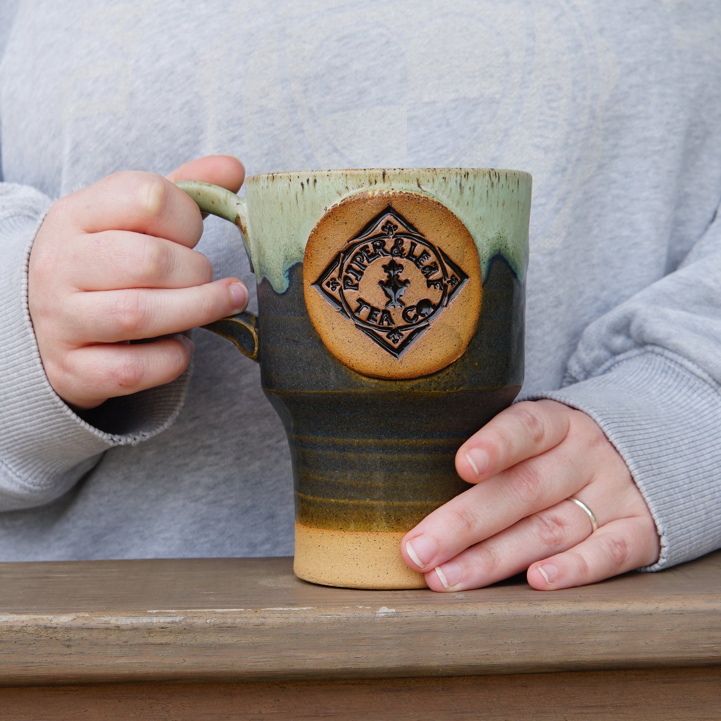 A person wearing a gray shirt holds a large Pottery Travel Mug with a Piper and Leaf Tea Co. logo while standing behind a wooden surface.