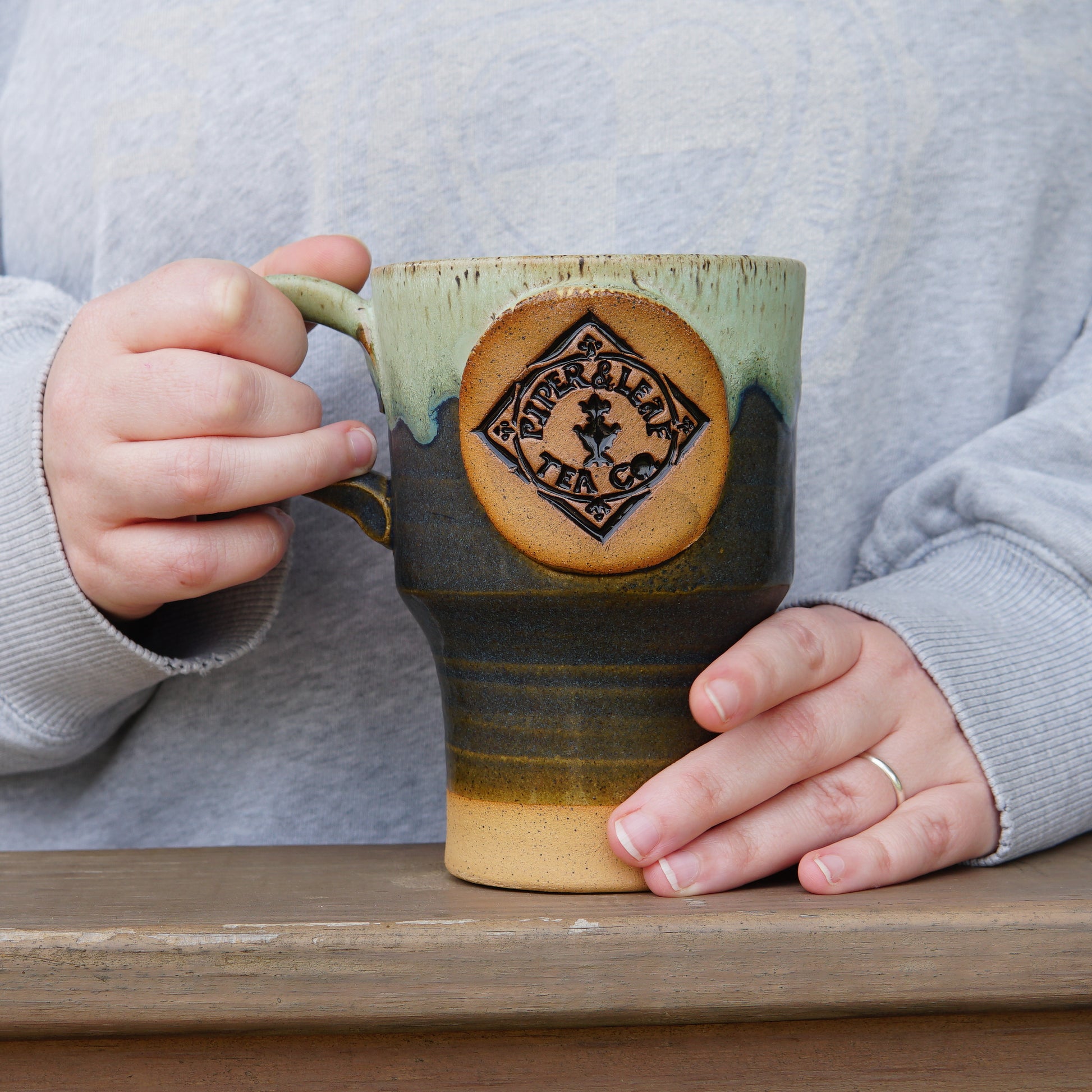 A person wearing a gray shirt holds a large Pottery Travel Mug with a Piper and Leaf Tea Co. logo while standing behind a wooden surface.