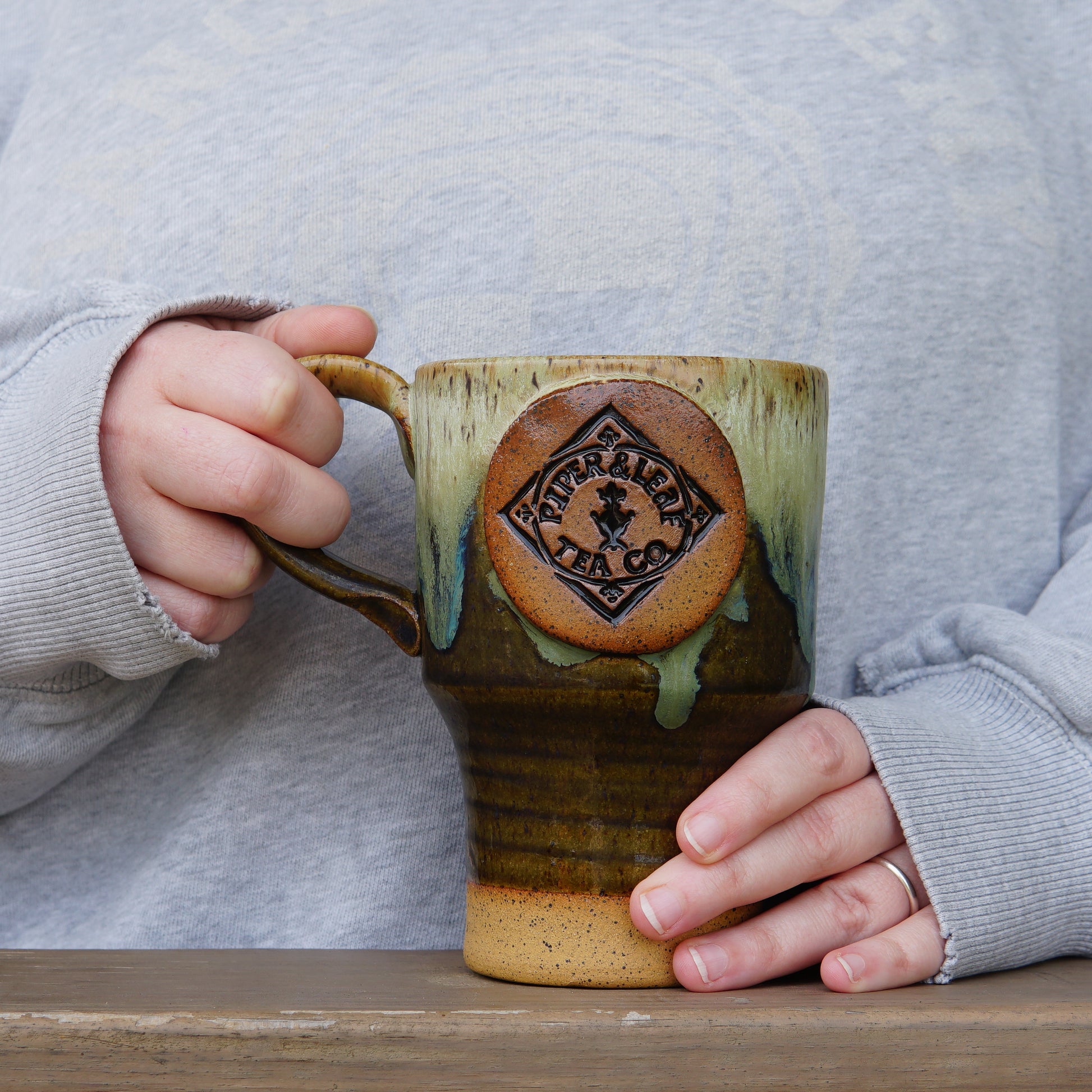 A person in a grey sweater holds a handmade Pottery Travel Mug from Piper and Leaf Tea Co., standing behind a wooden surface.