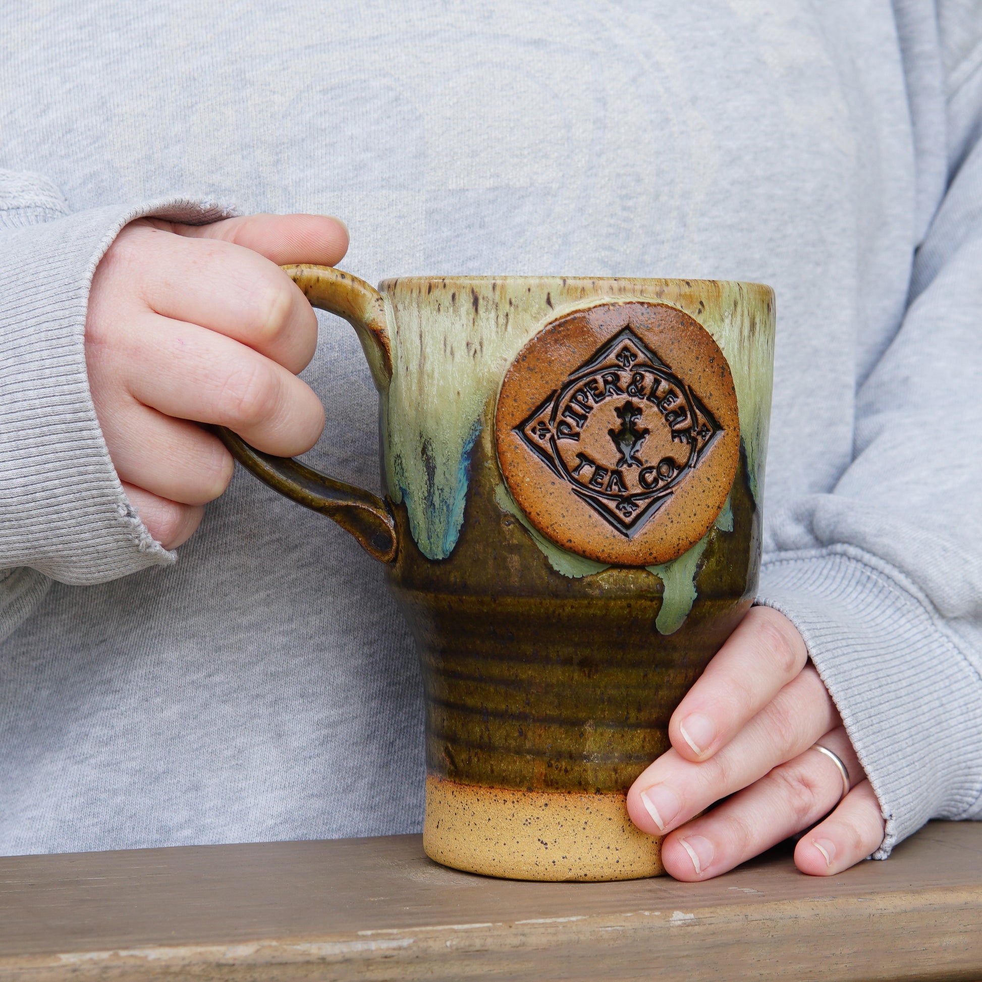 A person in a gray sweater holds a large, handcrafted Pottery Travel Mug with the “Piper and Leaf Tea Co.” logo, standing behind a wooden surface.