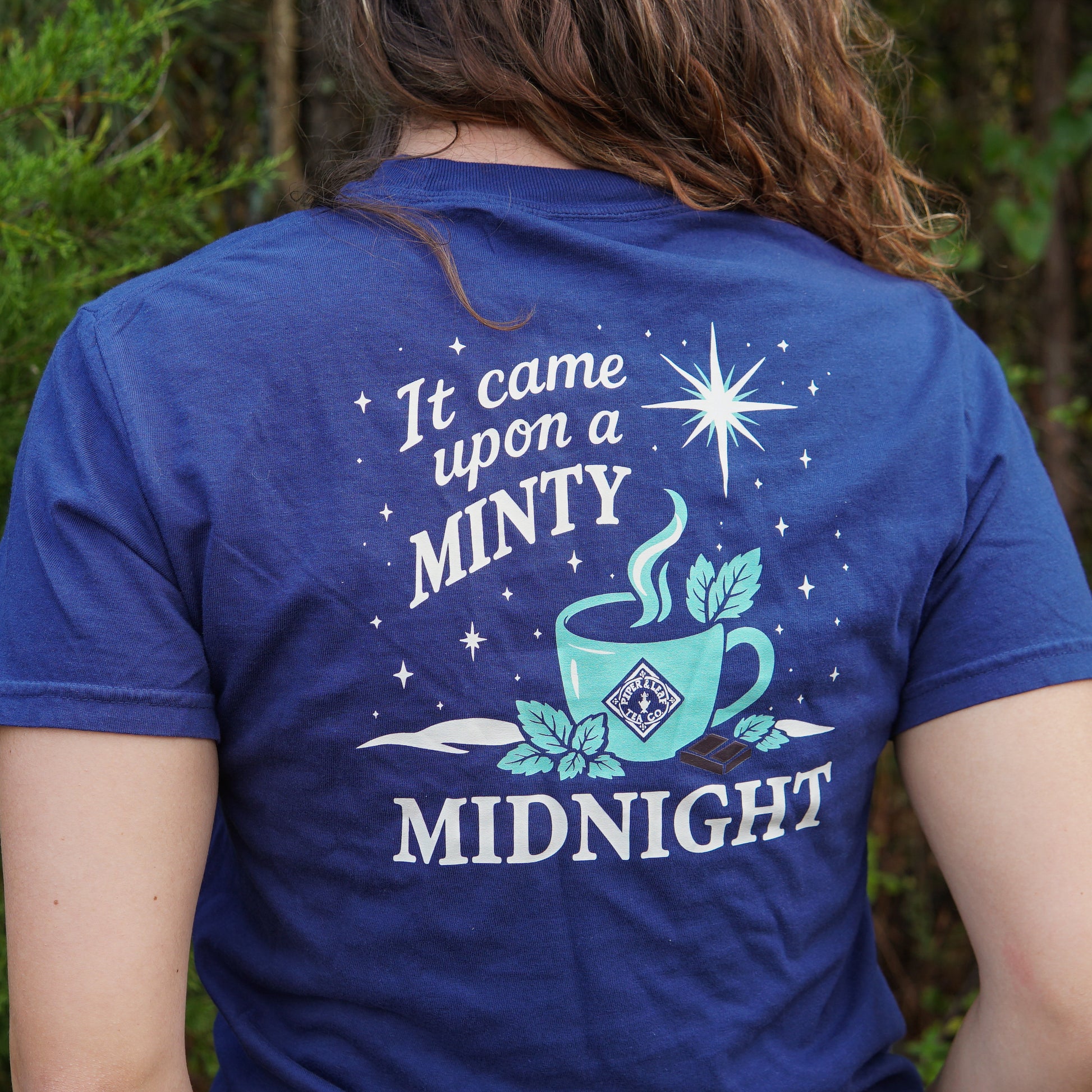 A person wears the Piper & Leaf Tea Co. "It Came Upon a Minty Midnight T-Shirt," featuring a steaming cup, stars, leaves, and the text "It came upon a MINTY MIDNIGHT" on the back.