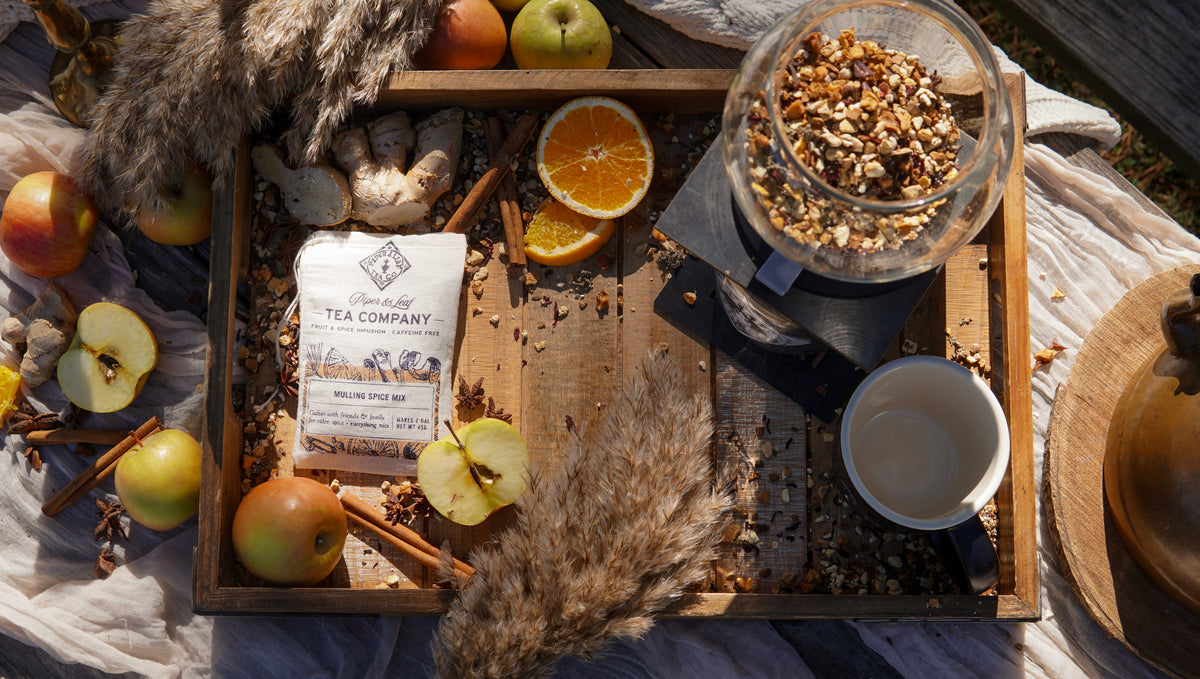 A wooden tray holds Piper & Leaf Tea Co.’s Mulling Spice Mix with a cup, glass jar of loose tea, fruits, cinnamon sticks, and dried flowers—ideal for serving festive drinks at holiday parties.