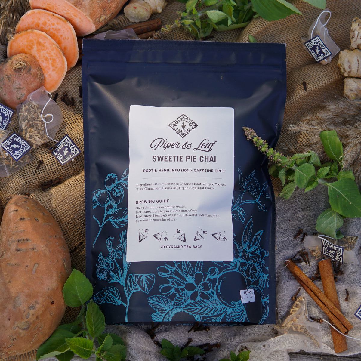 A package of Piper & Leaf Tea Co. Sweetie Pie Chai Bulk Sachets (70ct Tea Bags), a caffeine-free spiced herbal tea, is displayed with sweet potatoes, cinnamon sticks, ginger, tea bags, and fresh herbs on a rustic surface.