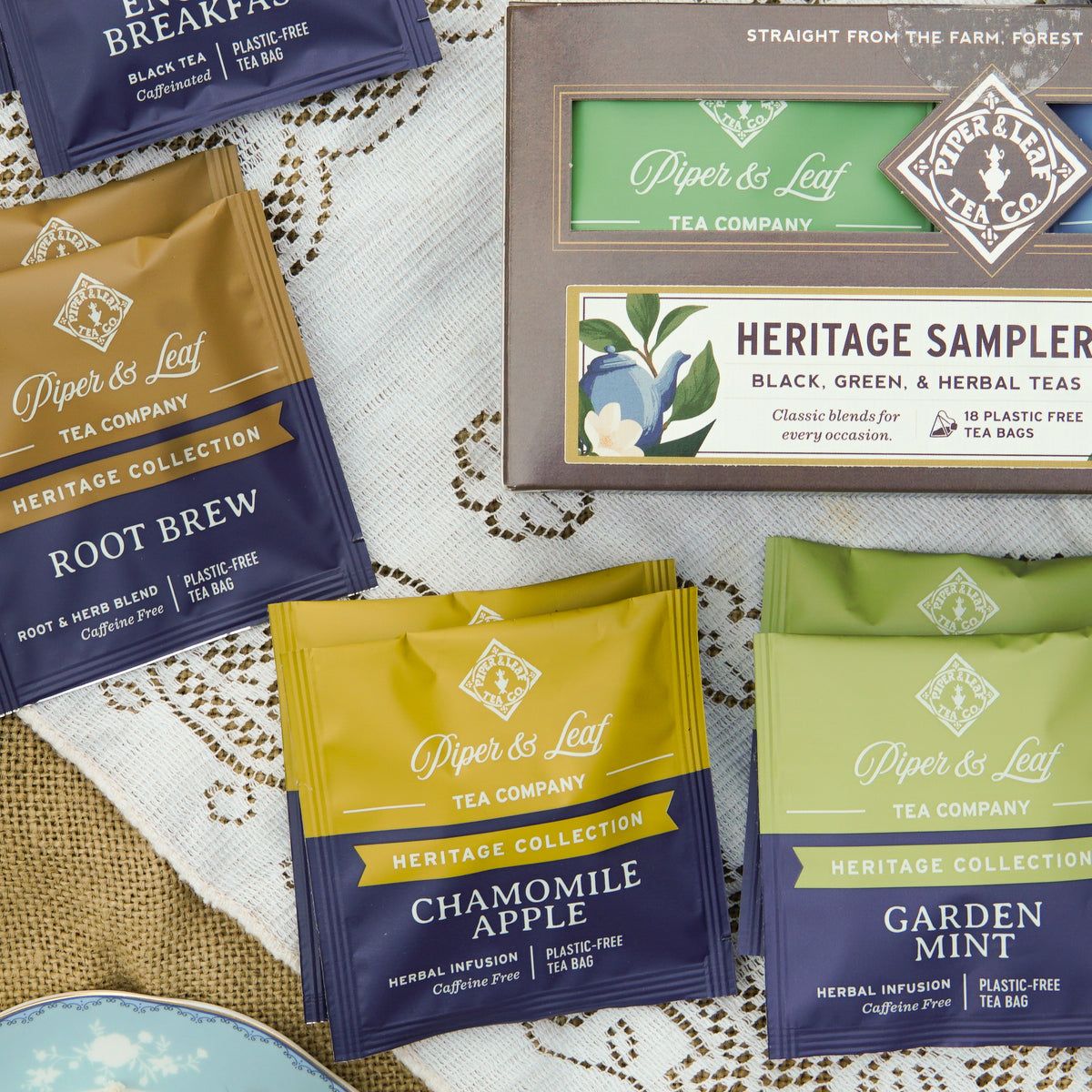 A selection of Piper and Leaf Tea Co.’s premium blends—Root Brew, Chamomile Apple, and Garden Mint—is artfully arranged on lace and burlap next to the elegant Heritage Sampler box of 18 tea bags.
